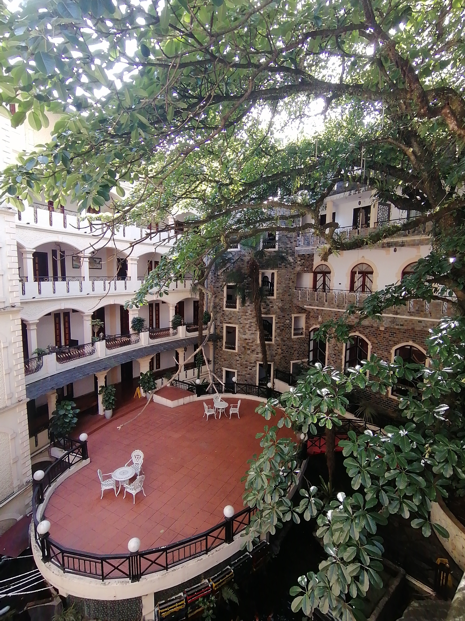 The image depicts an exterior view of a hotel courtyard in Sa Pa Ward, Vietnam. The courtyard is paved with red tiles and contains several sets of white patio furniture. A black wrought-iron railing surrounds a lower section of the courtyard. Two distinct architectural styles are present: one is a white facade with arched openings and decorative railings, and the other is constructed of stone with dark-framed windows. A large, mature tree with dense foliage overhangs the courtyard, casting shadows and filtering the sunlight. There are no people visible in the image.