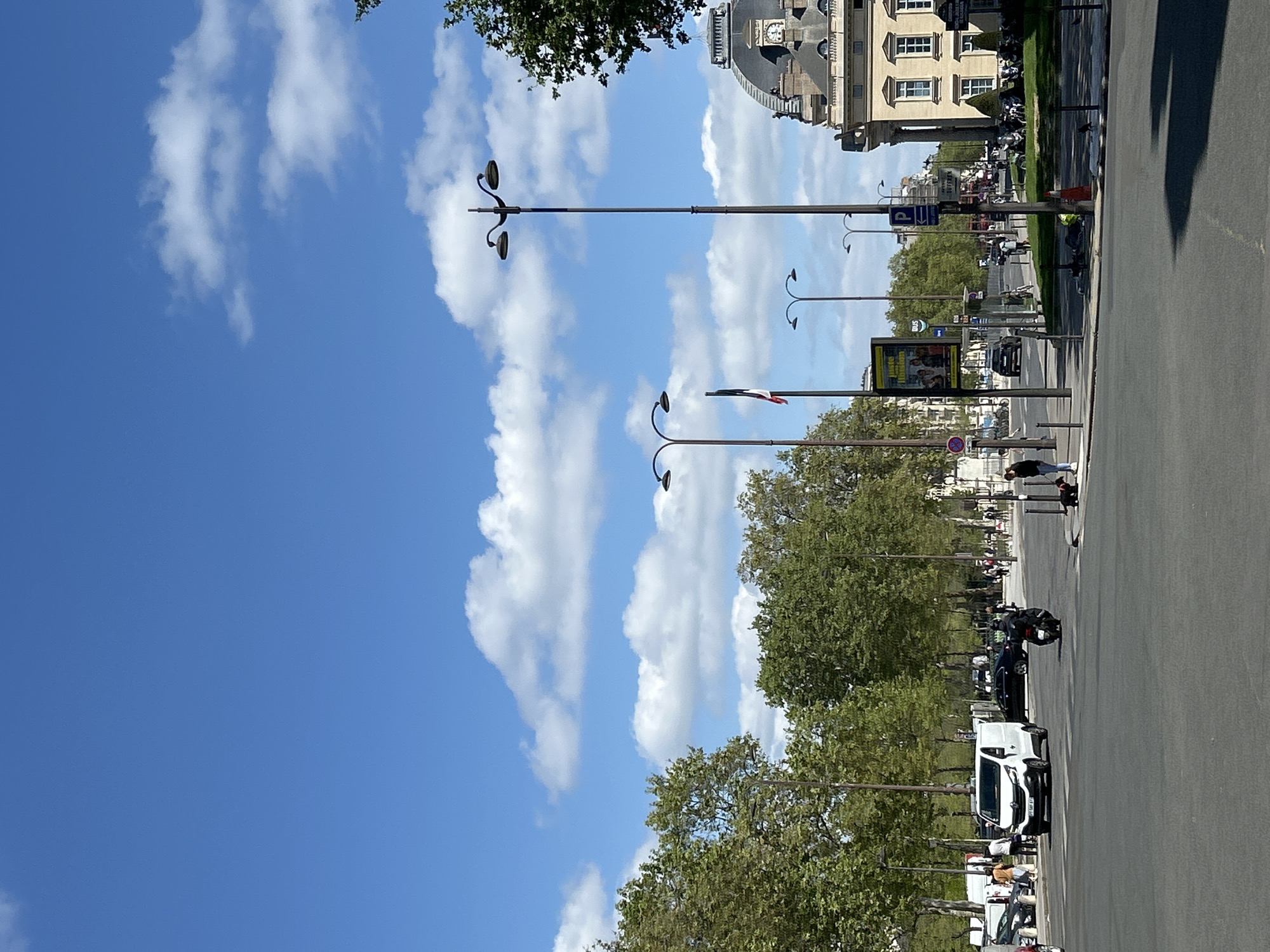 The image depicts a vibrant daytime street scene, likely in a European city given the architecture and street furniture. The perspective is from street level, looking down a wide boulevard lined with trees and buildings. The sky above is a clear blue adorned with scattered white cumulus clouds, suggesting fair weather.
In the foreground and midground, a broad asphalt road is visible, on which a white van, a black car, and a motorcycle are present. Multiple ornate, dark-colored streetlights with double arms line the street, casting distinct shadows on the pavement, indicative of bright sunshine. A prominent line of green trees runs along the length of the boulevard, separating the road from a pedestrian area or park, where several people are seen walking. One person in a dark top and light pants is walking on the sidewalk closer to the viewer.
In the background, grand, classical-style buildings dominate the skyline. One notable structure features a large domed roof with a clock tower. Other buildings display multiple windows and classical architectural elements. Among the street furniture, a yellow billboard poster is attached to a lamppost, with partially visible text that appears to read "Musées." A blue sign with a white "P" and an arrow, indicating parking, is also visible. A small flag, resembling the French tricolor (blue, white, red), is attached to one of the streetlights, serving as a strong location cue suggesting France. The white van has "FR" visible on its license plate. The overall impression is a bustling urban environment on a sunny day.