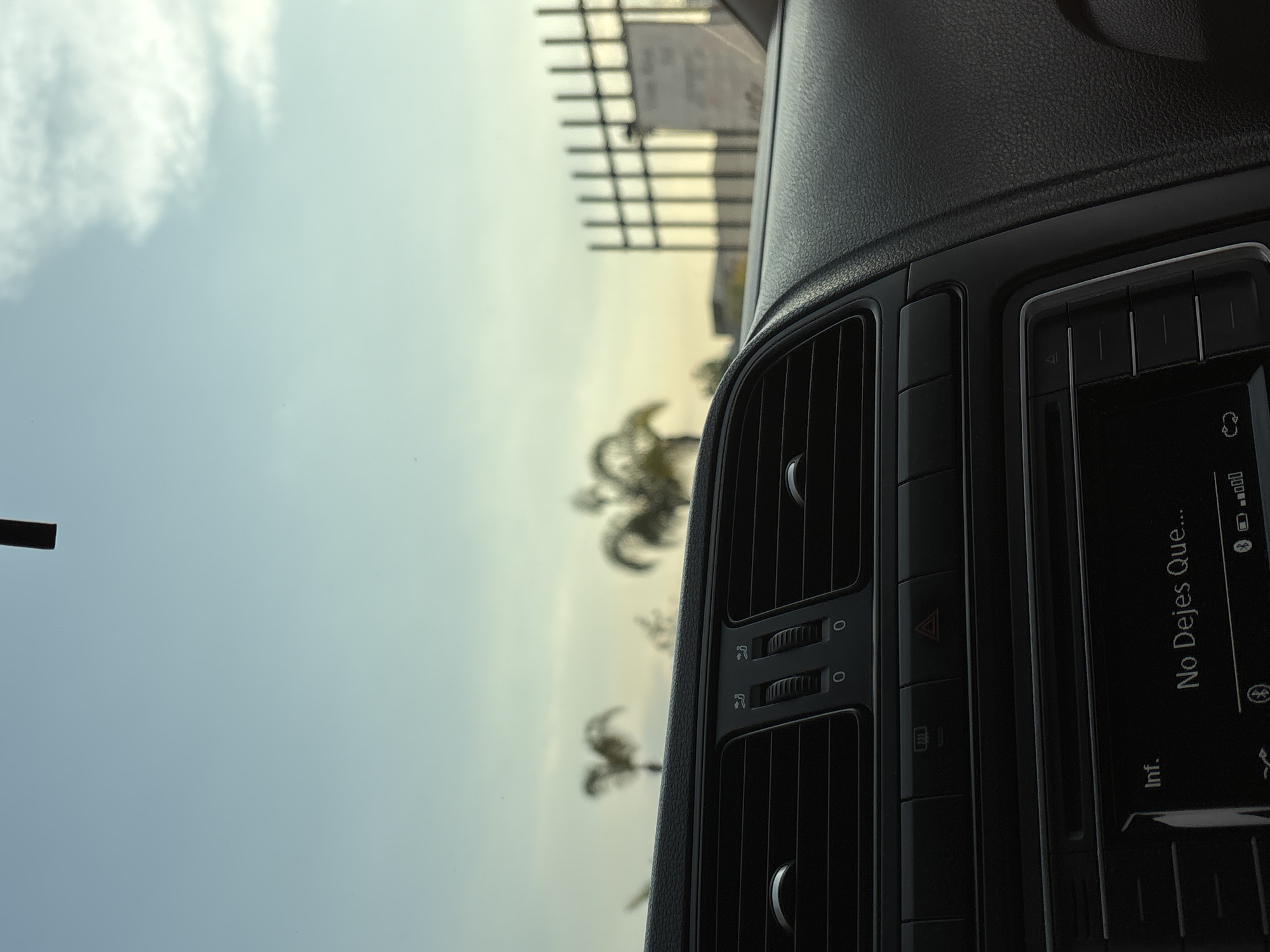 The image is taken from inside a vehicle, likely the driver's seat, focusing on the dashboard's central console. The console includes air vents and a car radio, displaying the message "No Dejes Que..." in Spanish. Through the windshield, the outside environment is visible. Palm trees are silhouetted against a bright, hazy sky, suggesting a sunny day. Faintly visible in the distance is a metal fence or structure. Based on the location provided, San Bernardino Tlaxcalancingo, Mexico, the vegetation is consistent with a subtropical climate. No people are visible in the frame. The scene depicts a stationary vehicle with a view of a typical day in that region.