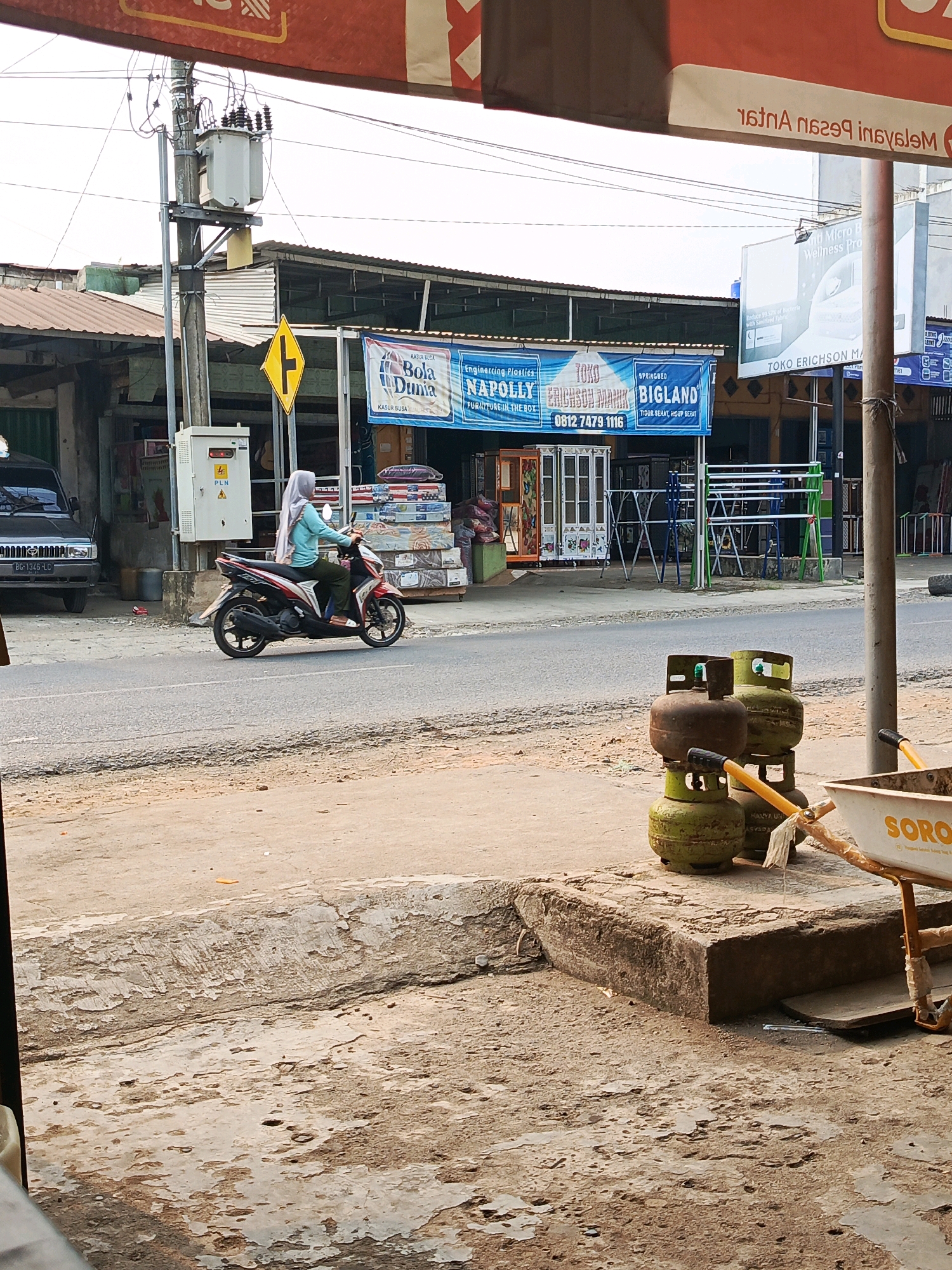 This is an eye-level, medium shot of a street scene in Megang Sakti II, Indonesia, likely during daytime. In the foreground, a concrete slab with a rough, textured surface leads to a pathway. To the right, a white wheelbarrow with orange handles and a yellow stripe bearing the word "SORO" is partially visible. Behind the wheelbarrow and on the concrete slab are several green propane gas tanks, some stacked.
In the midground, a woman wearing a light blue hijab and tunic, and dark green pants, is riding a red and white motorcycle with her right leg resting on the footboard. The motorcycle is facing left. The woman is looking forward and slightly to her left. A road runs horizontally across the image, with a sandy shoulder on the left and a paved surface where the motorcycle is.
Behind the road, there are several shops and buildings. To the left, a dark-colored SUV is parked. A utility pole with electrical transformers and wires stands prominently on the left side of the image. Yellow and black signs are visible, including one with a T-junction symbol and another with the "PLN" logo on an electrical box.
Further back, a banner is stretched across the front of a building, advertising "KASUR BUSA" (foam mattress) with brands like "Bola Dunia," "Napolly," and "Bigland." Various items, possibly mattresses and furniture, are displayed and stacked in front of the shops. The overall environment appears to be a commercial area with a mix of shops and street infrastructure. The sky is overcast.