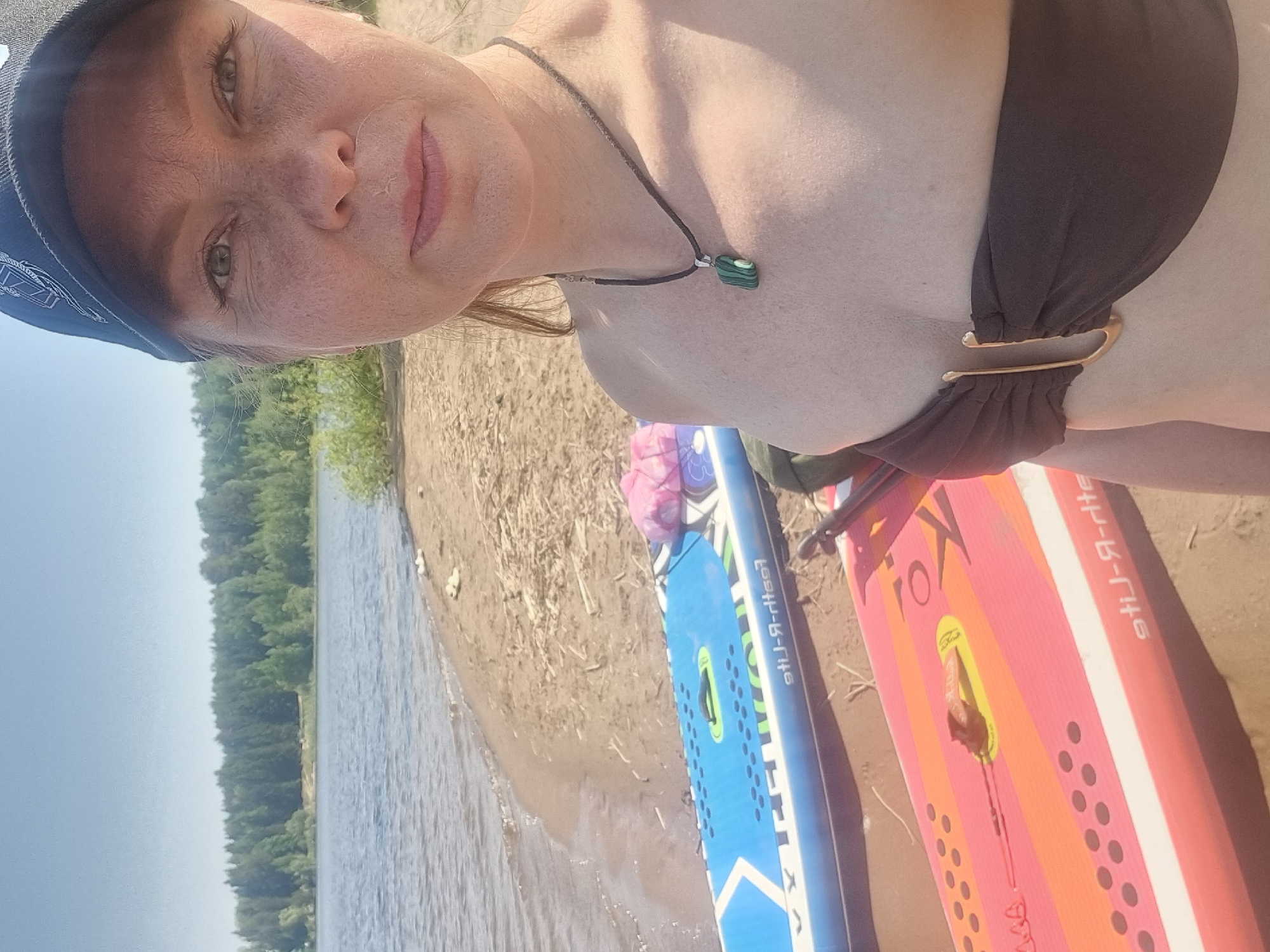 A woman with freckles on her face, wearing a black bikini top and a dark blue cap, is posing for a selfie on a beach. She is wearing a necklace with a green stone pendant. Two paddleboards, one blue and one pink, are visible in the lower part of the frame next to her. The beach is sandy, and the water is visible in the background, with trees lining the opposite shore. The location is likely near a body of water in Chaikovsky, Russia.