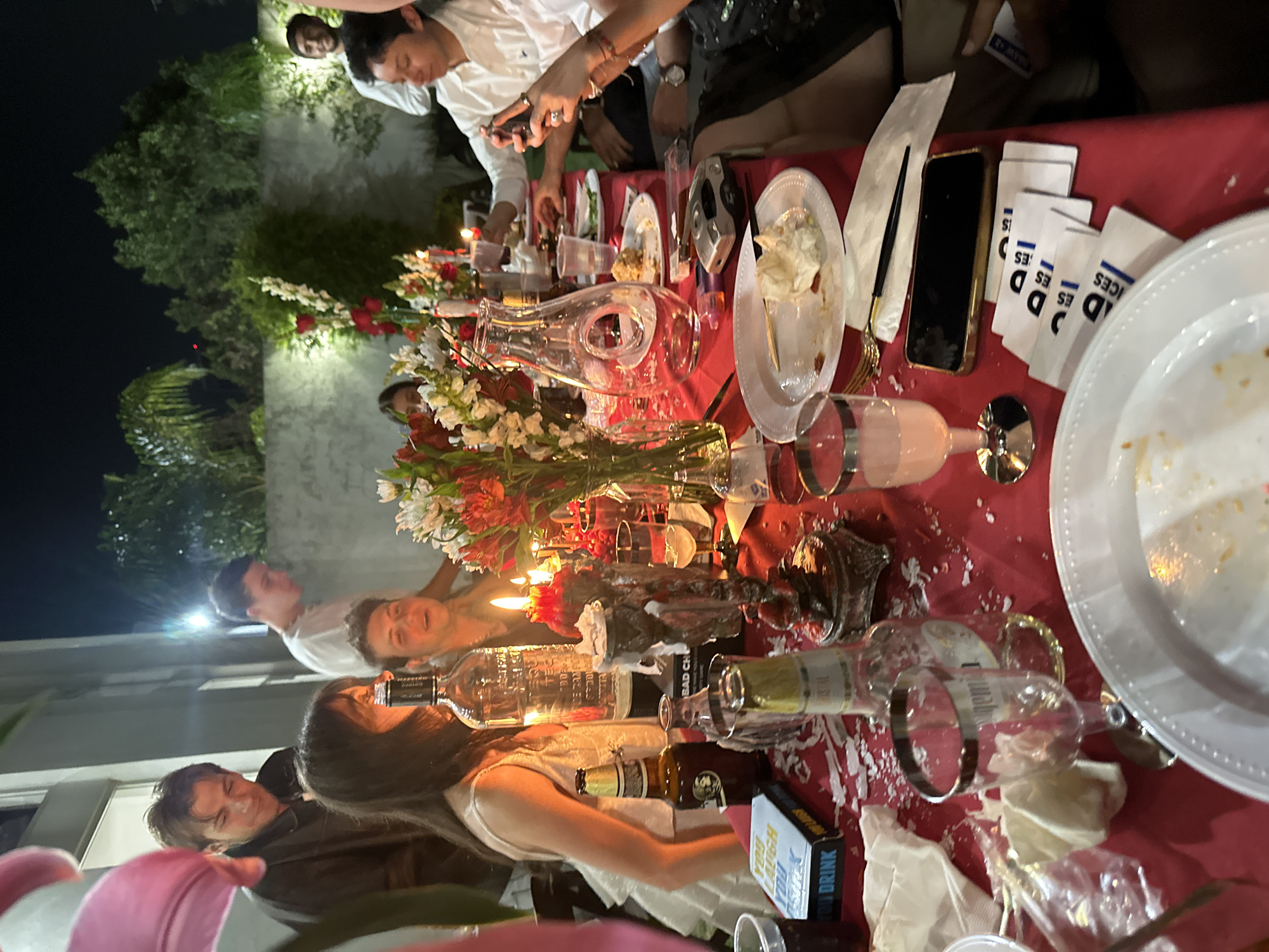 The image depicts a scene from an event, possibly a celebration, taking place outdoors, likely at night. The location appears to be Huixquilucan de Degollado, Mexico. A long table covered with a red tablecloth is heavily laden with plates, glassware, bottles of alcohol (including whiskey), flowers in vases, and scattered food. Several people are around the table, some interacting with each other, and one person taking a photo with a smartphone. There are candles, suggesting a festive or celebratory atmosphere. There are also scattered napkins and what appear to be small promotional cards or items with the text "ICES" or "DD ES" on them. One box is visible with "YOU LAUGH YOU DRINK" written on it. The environment shows trees and a building exterior.