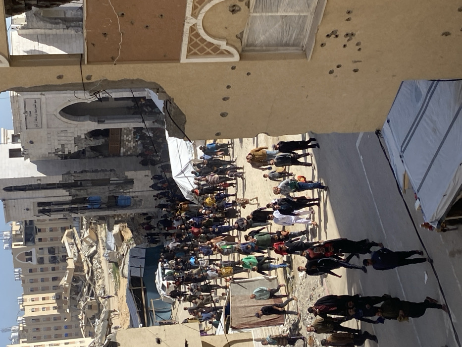 The image captures a densely populated street in Khan Yunis, Palestinian Territory, surrounded by extensive structural damage. In the immediate foreground on the right, a multi-story building with a light brown facade is visibly pockmarked with numerous circular impact points, consistent with bullet or shrapnel damage, and features a partially obscured or damaged window. A section of its upper right corner is missing, revealing internal elements.
The street below is crowded with a large congregation of people, including adults and children, moving in various directions, though primarily appearing to walk towards the background. Individuals are dressed in diverse civilian attire. Along the street, white canvas tents are erected on both the left and right sides, indicating temporary structures.
In the midground, a prominent, multi-story building with an arched entrance, displaying Arabic script above it, exhibits severe structural damage, including significant portions of its facade collapsed and internal elements exposed. To its left, a vast area is covered by large piles of rubble and debris, signifying widespread destruction of structures. Beyond this, multi-story residential buildings are visible, some appearing damaged, others partially intact under a clear sky.