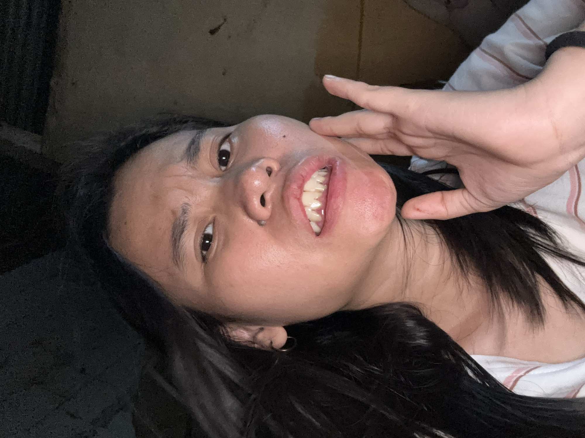 A young woman is captured in a close-up, front-facing selfie, her face angled slightly to the right. She has long, dark hair that is partially obscuring her left cheek and shoulder. Her mouth is open, revealing her teeth, and her expression is one of playful exaggeration or discomfort. Her right hand is raised and touching her cheek, with her fingers splayed. She is wearing a striped shirt, and a silver hoop earring is visible in her left ear. The background is dark and appears to be an indoor setting, possibly a simple dwelling with a corrugated metal ceiling visible at the top of the frame. The lighting is dim, creating shadows on her face and the background. The overall impression is of an informal, personal moment captured in what seems to be a modest environment within East Jakarta, Indonesia.