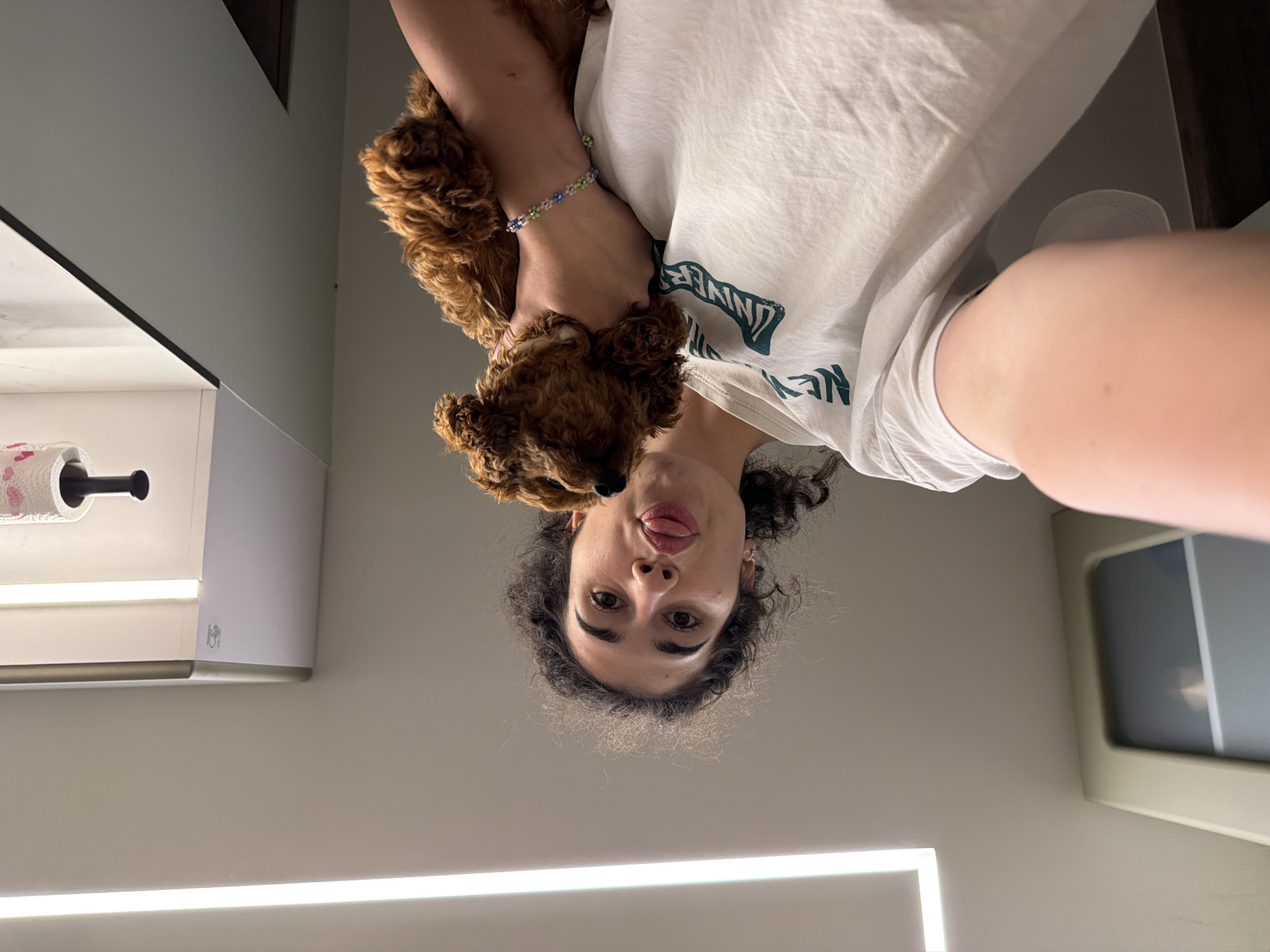 The image, taken from a low angle, shows a young woman with curly dark hair and a white t-shirt holding a small, reddish-brown poodle puppy on her shoulder. She is sticking her tongue out playfully. The puppy is resting on her arm, which is adorned with a beaded bracelet featuring small flowers in blue, green, and white. In the background, a bright vertical light fixture is visible on the left, and a roll of toilet paper with a heart pattern is mounted on the wall above. The environment appears to be indoors, possibly a kitchen or bathroom, with modern cabinetry and lighting. The overall scene is casual and lighthearted. There is no information in the image to suggest the location is Sofia, Bulgaria.