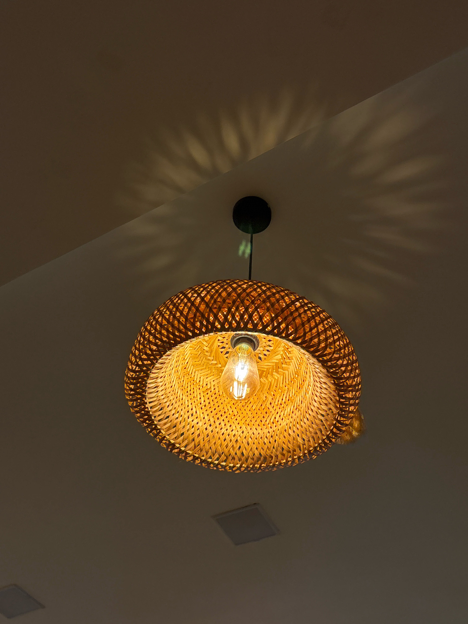 A woven bamboo pendant light hangs from a dark ceiling, casting a patterned shadow. A lit Edison bulb is visible within the shade. Two recessed square ceiling fixtures are also present. The scene is indoors, on a ceiling.