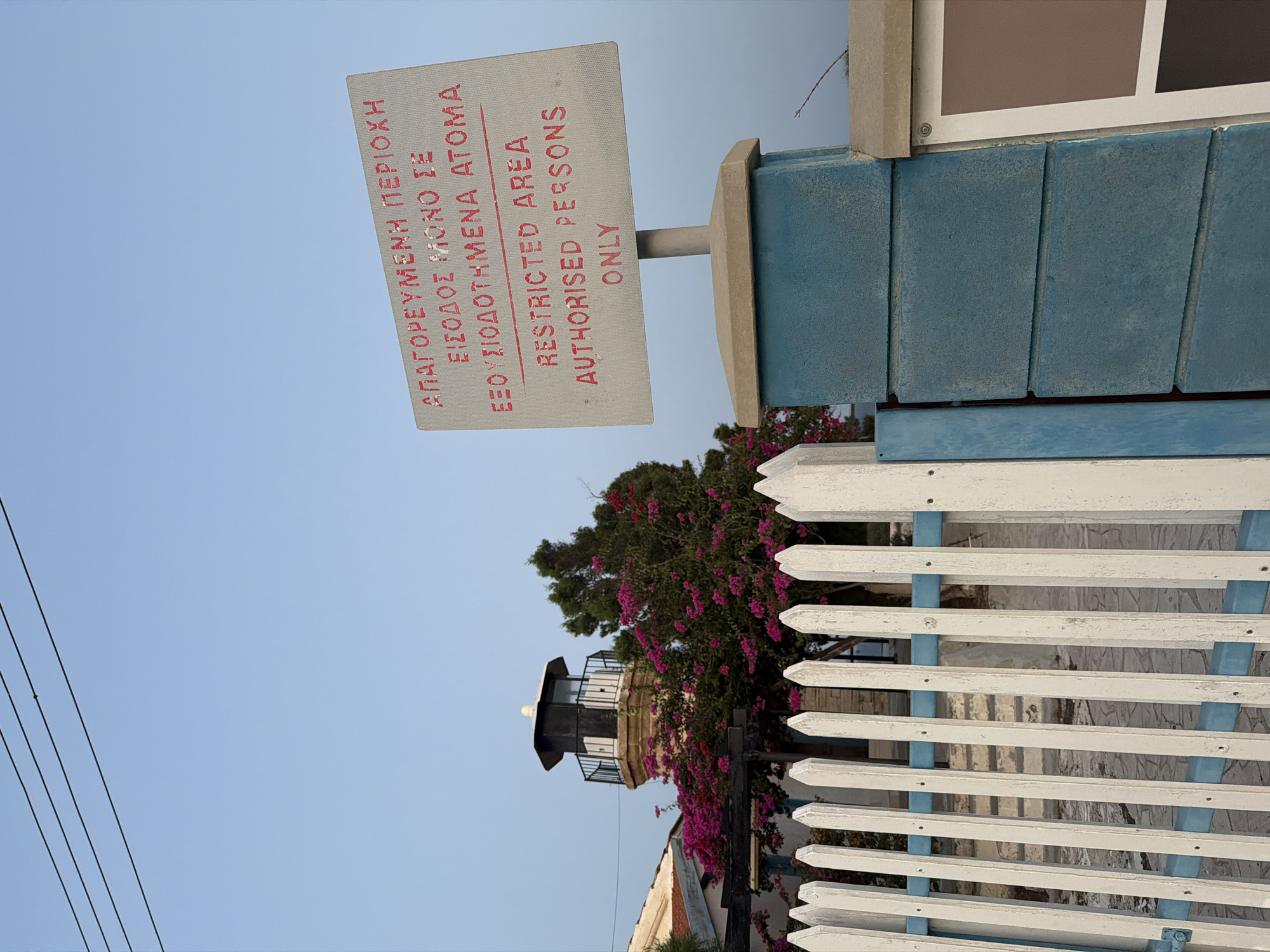 The image shows a signpost in front of a white picket fence and a lighthouse-like structure. The signpost has two sections of text. The top section is in Greek, and the bottom section is in English. The Greek text reads "ΑΠΑΓΟΡΕΥΜΕΝΗ ΠΕΡΙΟΧΗ ΕΙΣΟΔΟΣ ΜΟΝΟ ΣΕ ΕΞΟΥΣΙΟΔΟΤΗΜΕΝΑ ΑΤΟΜΑ" which translates to "Restricted Area Entrance only for authorized persons". The English text below reads "RESTRICTED AREA AUTHORISED PERSONS ONLY". The sign is attached to a blue concrete post, which is cemented into a beige rectangular base. The white picket fence has a blue horizontal bar running through it. In the background, a lighthouse-like structure with a dark top is visible, surrounded by bright pink bougainvillea flowers and green foliage. The sky is clear and light blue, with a few black electrical wires visible in the upper left corner. The overall scene suggests a private or secure area, possibly in a coastal town given the presence of the lighthouse.
