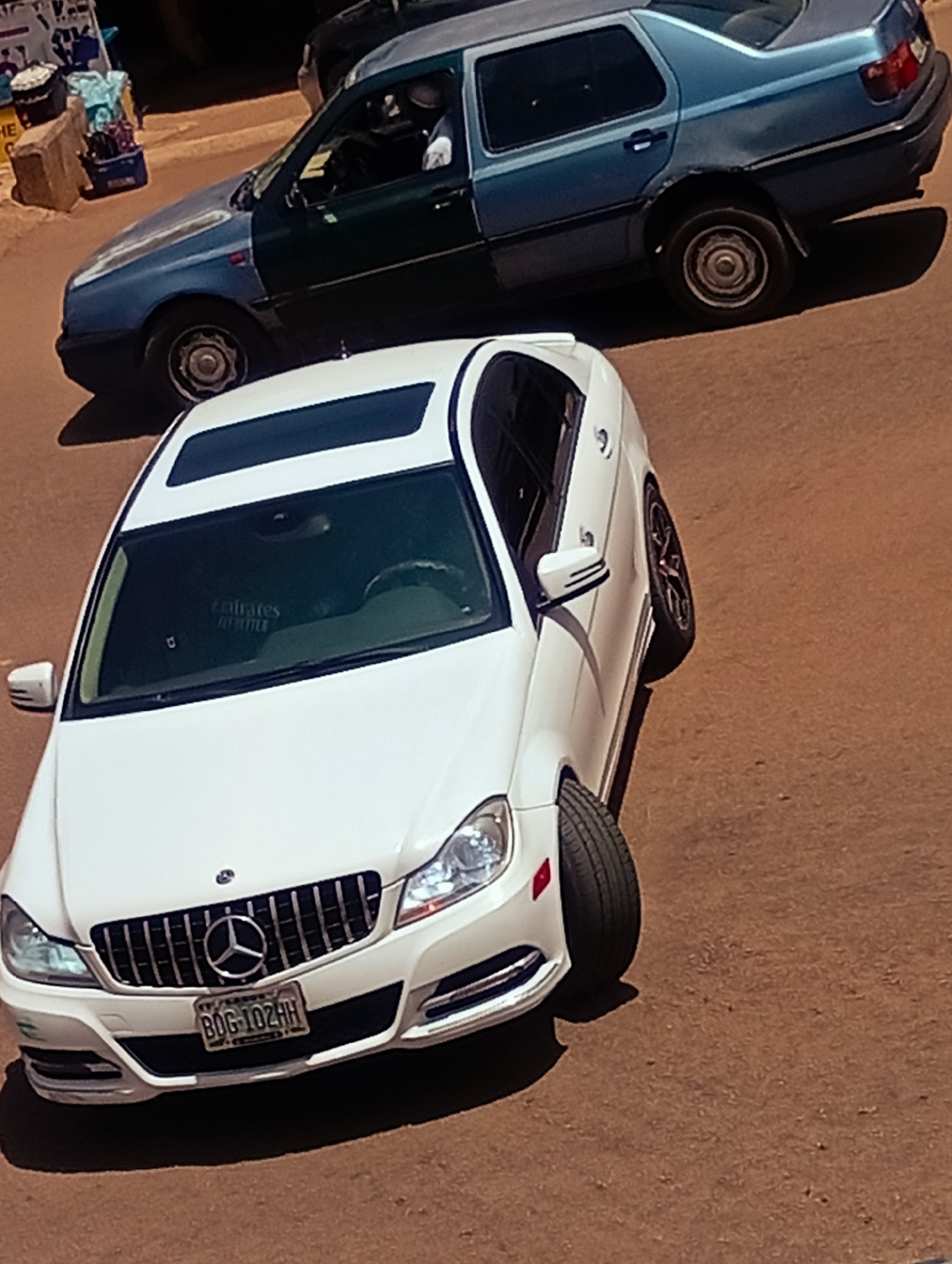 The image shows a street scene in Bukuru, Nigeria, featuring two cars. In the foreground, a white Mercedes-Benz sedan is parked, facing left. It has a sunroof, a distinctive grille with a Mercedes-Benz logo, and a license plate that reads "BDG-102HH". The car's front passenger side tire is turned outwards. Behind the Mercedes, a blue and green Volkswagen sedan is visible, also parked. The Volkswagen appears to have some damage to its rear quarter panel. The ground is a reddish-brown dirt. In the upper left corner of the image, there is a blurry view of a street stall with various items, including bags and containers. The overall lighting suggests it is daytime.