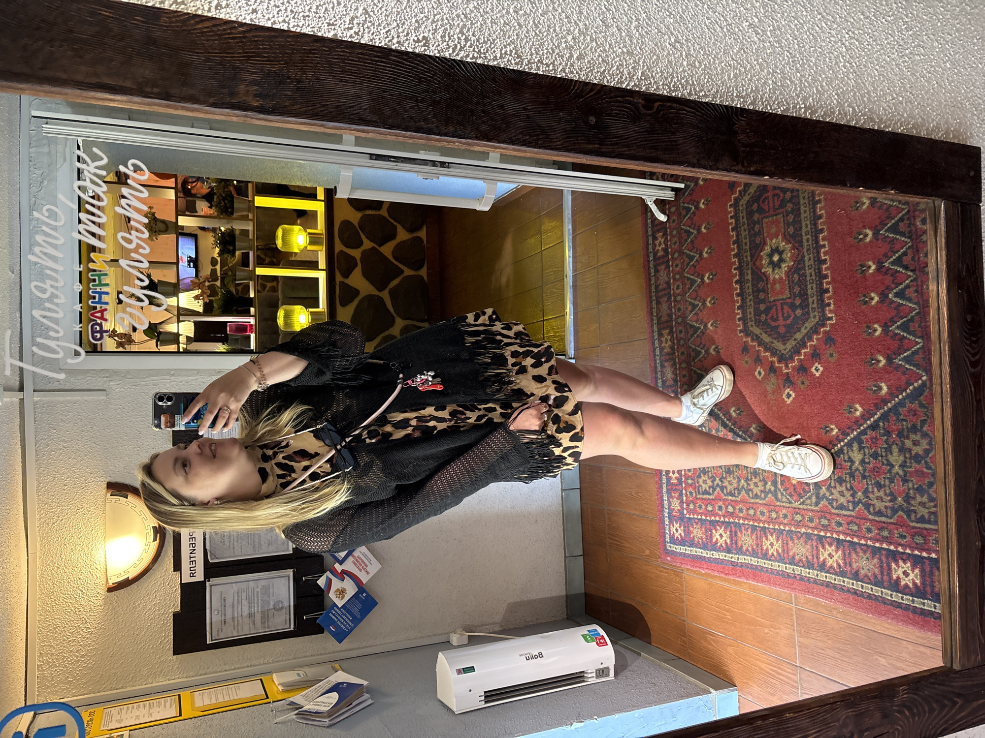A woman is taking a mirror selfie in what appears to be the entrance area of a cafe or restaurant, likely located in Chaikovsky, Russia. The woman is blonde, wearing a leopard print dress with a black open-knit cardigan with fringe detailing. She has a crossbody bag with a chain strap and sunglasses hanging from it. In the background, there's a bulletin board with documents, a white electric heater on the wall, and a large mirror reflecting the scene. Through an open doorway, the interior of the establishment is visible, featuring illuminated shelves with plants and decorative elements, with text on the glass door that reads "Гулять, Франц, пить, гулять" (meaning "Walk, Franz, drink, walk"). The floor is covered with wooden planks and a red oriental rug.