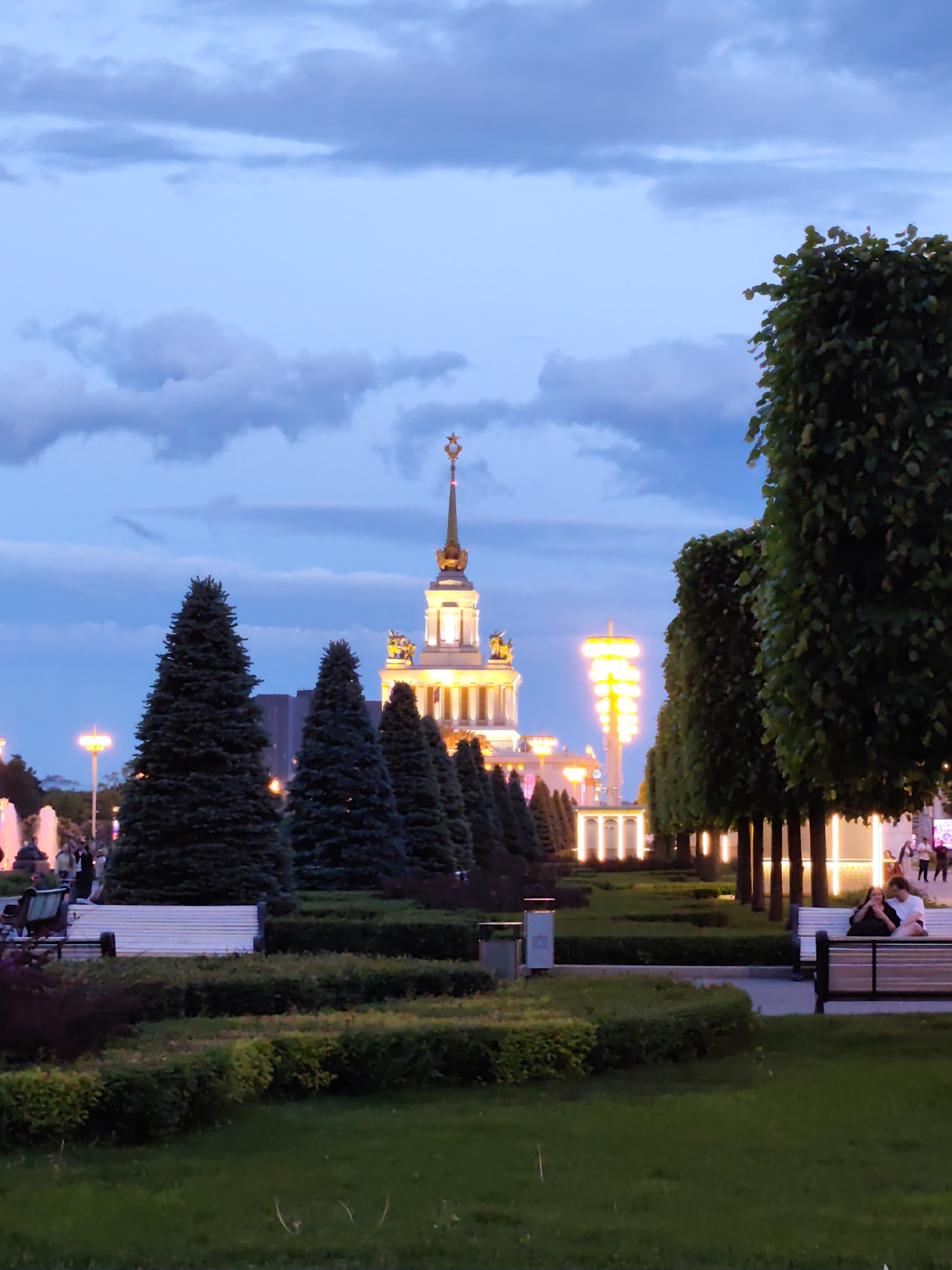 The image captures a twilight scene in Moscow, Russia. The prominent building in the background is illuminated, featuring a spire topped with a star. In the foreground, there is a park area with manicured lawns, shrubs, and rows of trees, some of which are evergreen. Benches are visible, and a couple is sitting on one. In the distance, there are fountains.