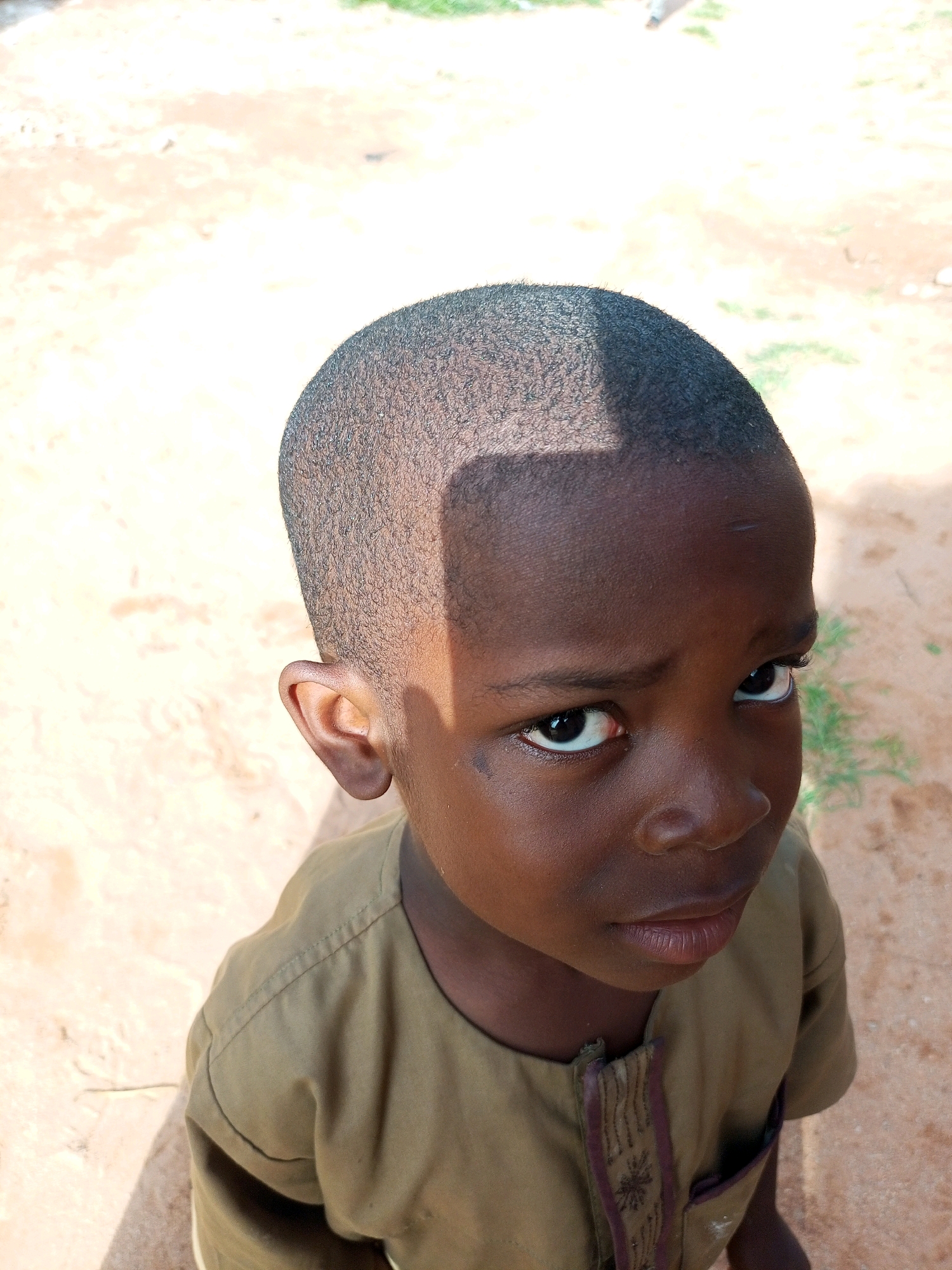 An overhead medium shot captures a young African boy standing outdoors. The boy is facing forward, looking slightly up and to his right with his head angled. He has short, dark, textured hair and a dark complexion. His expression is solemn, with his eyes open and looking directly at the camera. A faint shadow, likely from an overhead object, crosses his forehead diagonally from left to right. He is wearing a simple, light brown or olive-drab shirt with a darker, decorative trim running down the placket, featuring an embroidered star-like pattern. The shirt appears to have a small, open pocket on the right side.
The background consists of the ground, which is primarily sandy and light brown, with patches of dried grass and some sparse green vegetation. The lighting suggests a bright, sunny day, creating distinct shadows. The overall setting appears to be an outdoor, possibly rural or less developed, area. No specific landmarks or urban features are visible to indicate a precise location within Tempure, Nigeria. The image composition focuses tightly on the boy's face and upper body, with the environment serving as a blurred backdrop.
