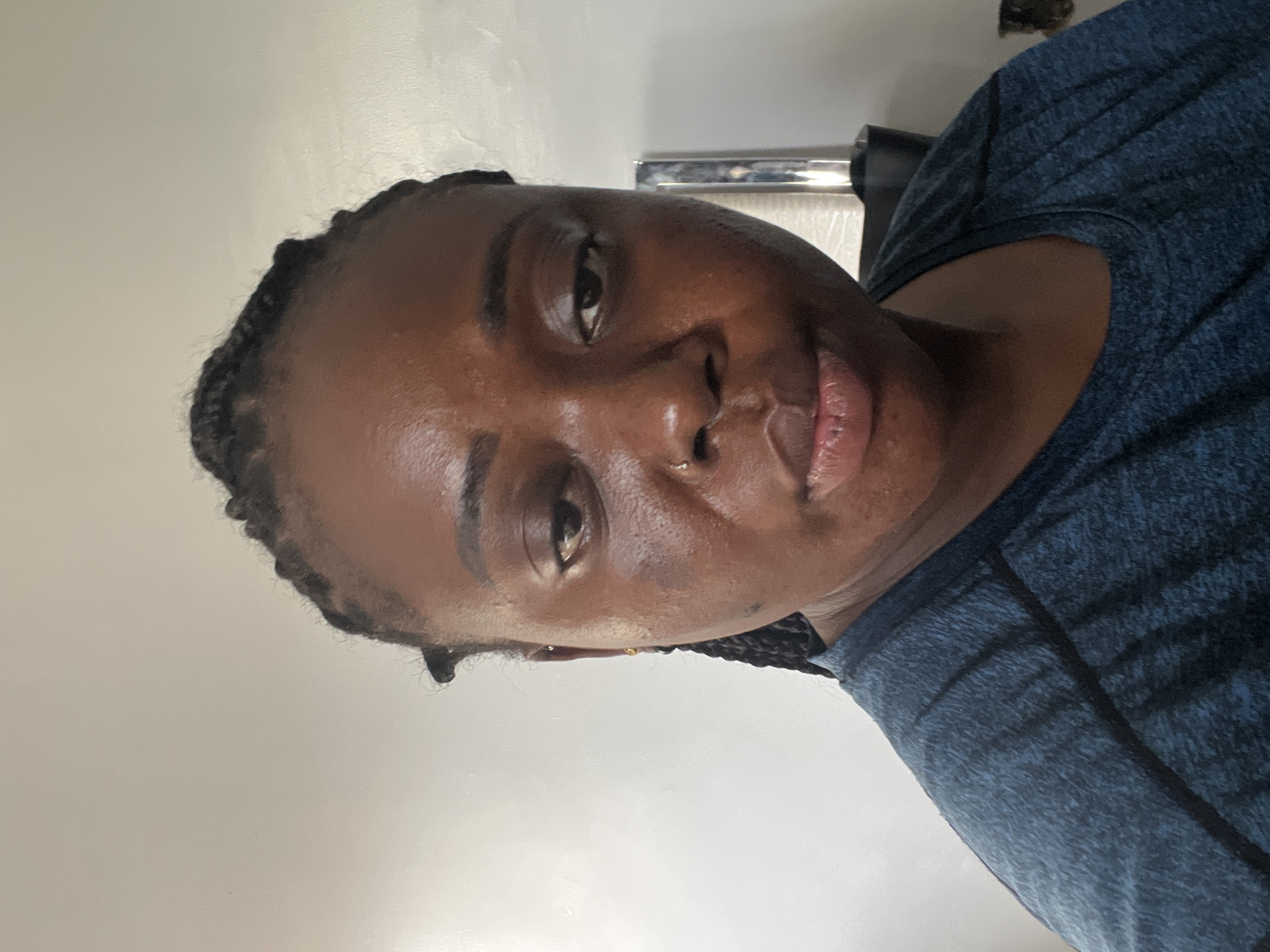 A close-up, tilted head-and-shoulders shot of a dark-skinned woman. She has dark hair styled in braids and is wearing a blue shirt with dark accents. The background is a plain, light-colored wall. There is a metal object visible behind her head. No specific details indicate the location is Kubwa, Nigeria.