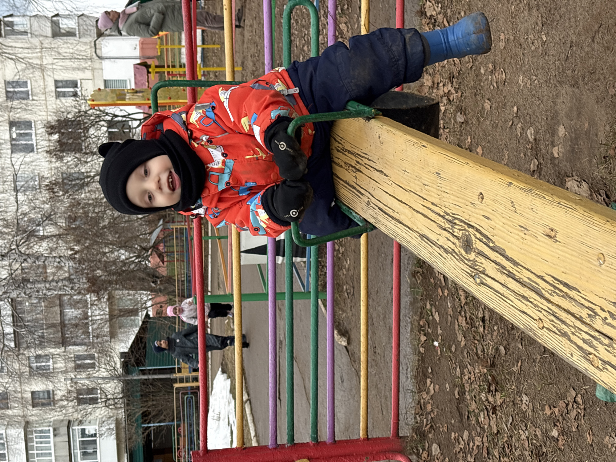A young child in a red coat, black hat, gloves, and blue boots is playing on a seesaw in a playground. The seesaw is made of wood and the playground has colorful metal bars. In the background, a multi-story building is visible. Other people are walking in the background. The ground is covered with dirt and leaves. The image was taken in the city of Chaikovsky, Russia.