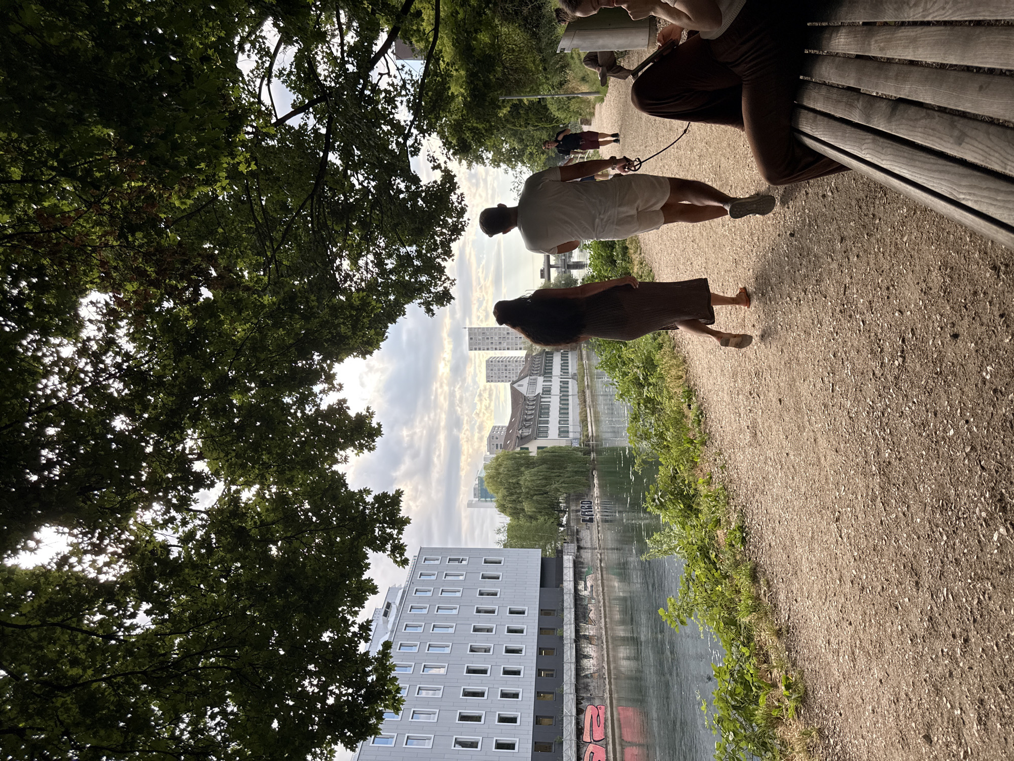 This is a vertical, eye-level, outdoor photograph of a riverside path in Zurich, Switzerland, under a canopy of large green trees. In the foreground, a wooden park bench is occupied by two individuals; one is seated and looking at a tablet, while the other's legs are visible in the lower right corner. The path is made of light brown gravel, with the river on the left, bordered by lush green vegetation and the side of a modern, grey building with rows of windows. Further along the path, a couple walks away from the camera, with the man holding a leash attached to a dog, which is not fully visible. The man is wearing a white t-shirt and white shorts, and the woman is wearing a long, brown dress. In the midground, across the river, a street with buildings and a large weeping willow tree is visible. In the background, more buildings and a bridge can be seen against a cloudy sky. The lighting suggests it is late afternoon or early evening.
