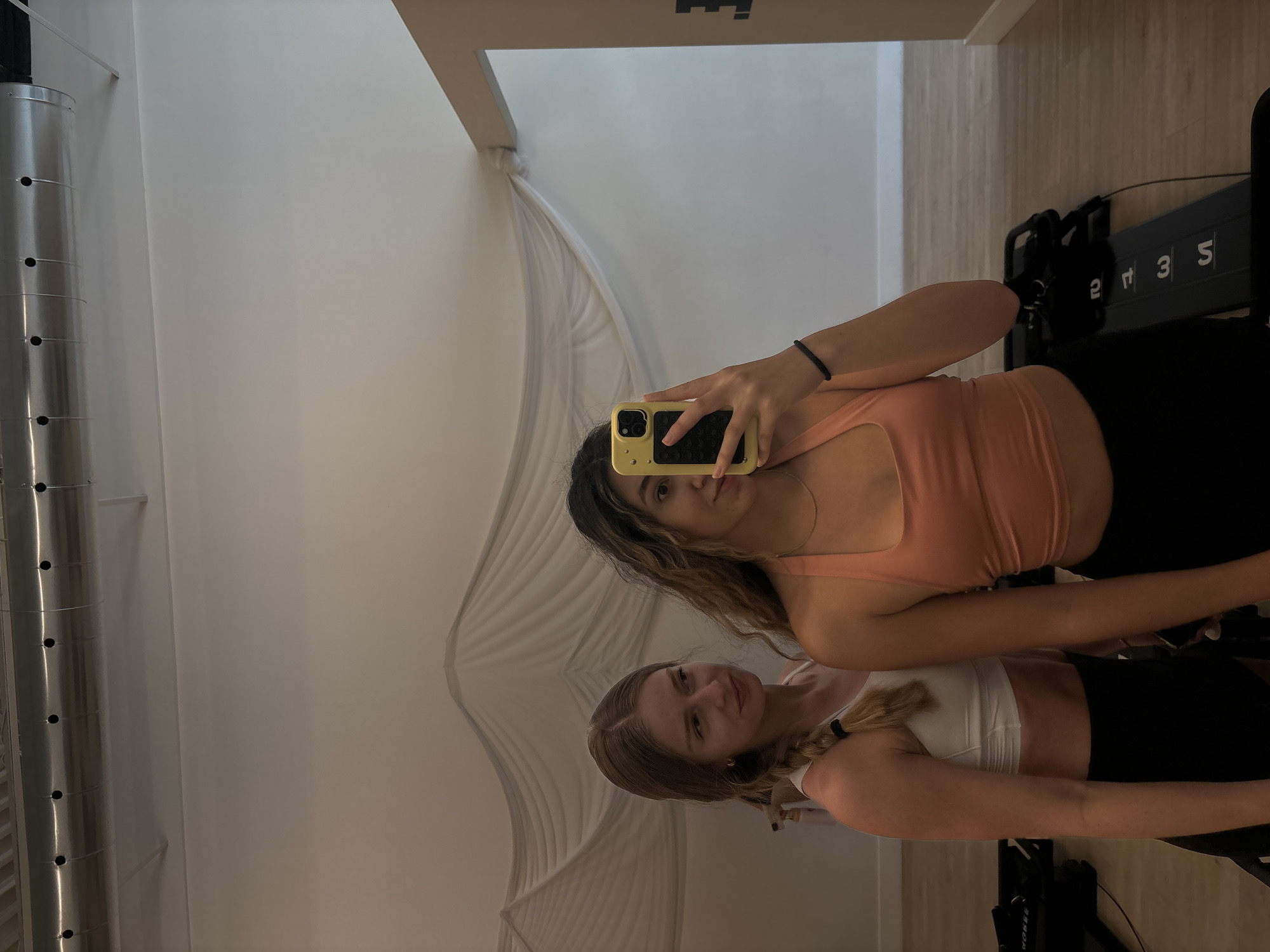 Two women are taking a selfie in front of a mirror inside what appears to be a fitness studio, potentially located in Delray Beach, Florida. The woman closer to the camera is holding a yellow-cased phone. She has brown hair and is wearing a peach-colored workout top and black bottoms. The second woman, reflected lower in the mirror, has blonde hair in a braid and is wearing a white top with black bottoms. In the background, a silver pipe with holes runs vertically, and a white, draped material is visible behind them. Exercise equipment, including a black machine with numbered increments, is also visible. The mirror reflects the scene, creating a layered perspective.