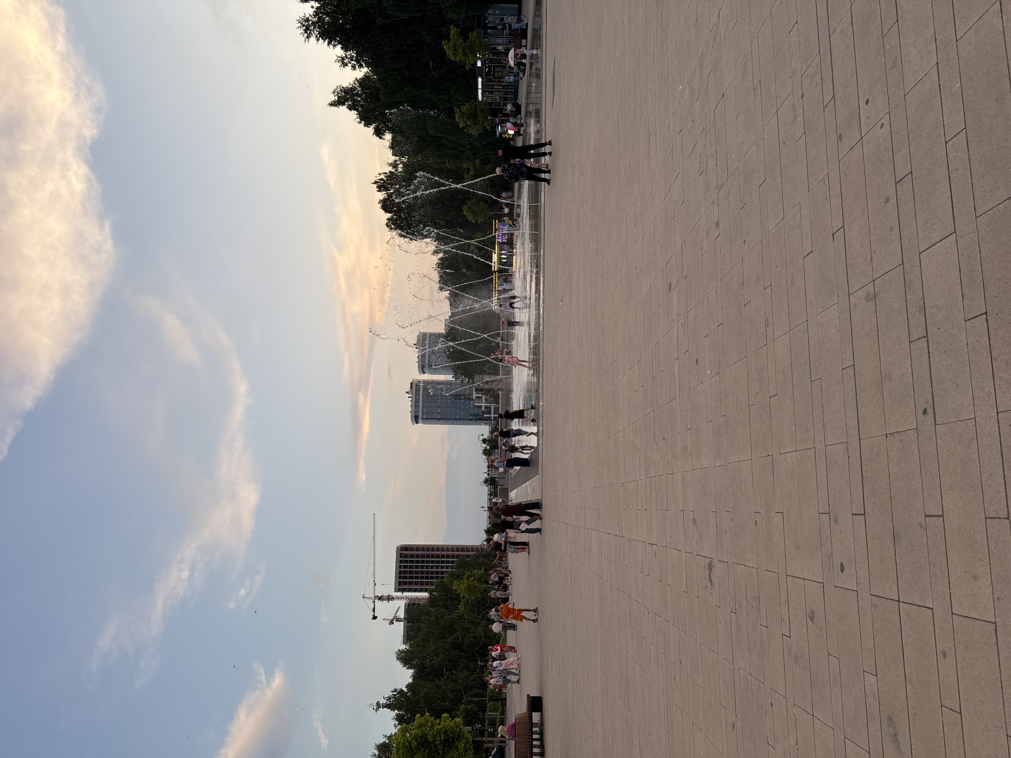 The image depicts a public space with a large paved area in the foreground, leading to a fountain and a group of people. In the background, there are trees, several modern buildings, and construction cranes. The sky is a soft blue with scattered clouds tinged with pink and orange, suggesting it is either early morning or late evening.
People are seen walking, standing, and interacting with the fountain. Some are children playing in the water, while others are adults observing or relaxing. The architecture suggests an urban environment, possibly a park or a plaza. The presence of construction cranes indicates ongoing development in the area.
This scene is located in the Izhevsk Central Park, specifically in the area of the central fountain. The fountain is a prominent feature, with water jets creating a dynamic display. The paved area is made of large, rectangular tiles. Trees line the perimeter of the park, and several cafes or small shops are visible near the fountain. The buildings in the background are modern high-rises, and the construction sites suggest urban expansion.