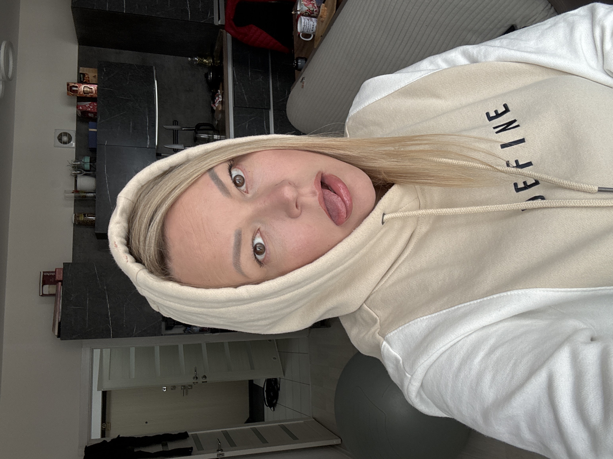 A woman with blonde hair, wearing a beige and white hoodie, is making a silly face with her tongue sticking out. She is indoors, likely in her apartment. In the background, there is a gray couch, kitchen cabinets, and a doorway leading to another room with shelves. A gray exercise ball is also visible. The specific location within Izhevsk, Russia is not discernible from the image.