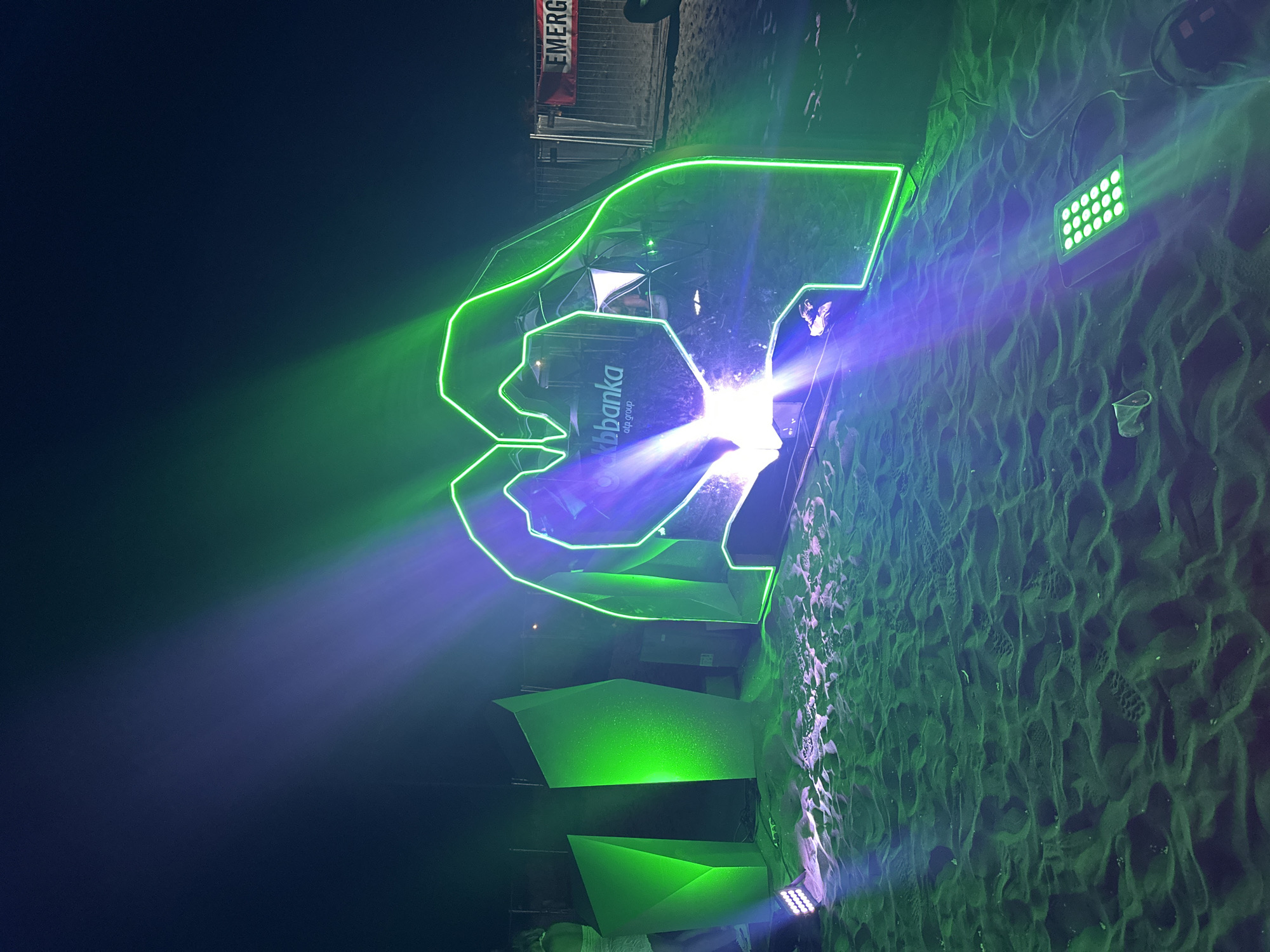 In Donji Štoj, Montenegro, a nighttime scene unfolds on a sandy beach, illuminated by vibrant neon lights. A large, stylized heart shape, outlined in bright green neon, dominates the center of the frame. Inside the heart, a logo for "kbbanka atp group" is visible. To the left of the heart are several angular, green illuminated structures. The sand is uneven, marked with footprints and scattered with a few small objects, possibly debris. Several green LED spotlights are positioned on the sand, casting beams of light that interact with the neon glow, creating a dramatic atmosphere. In the background, a fence is visible, with a red banner displaying the word "EMERGENCY" attached to it. The overall impression is an outdoor event or installation at dusk or night.