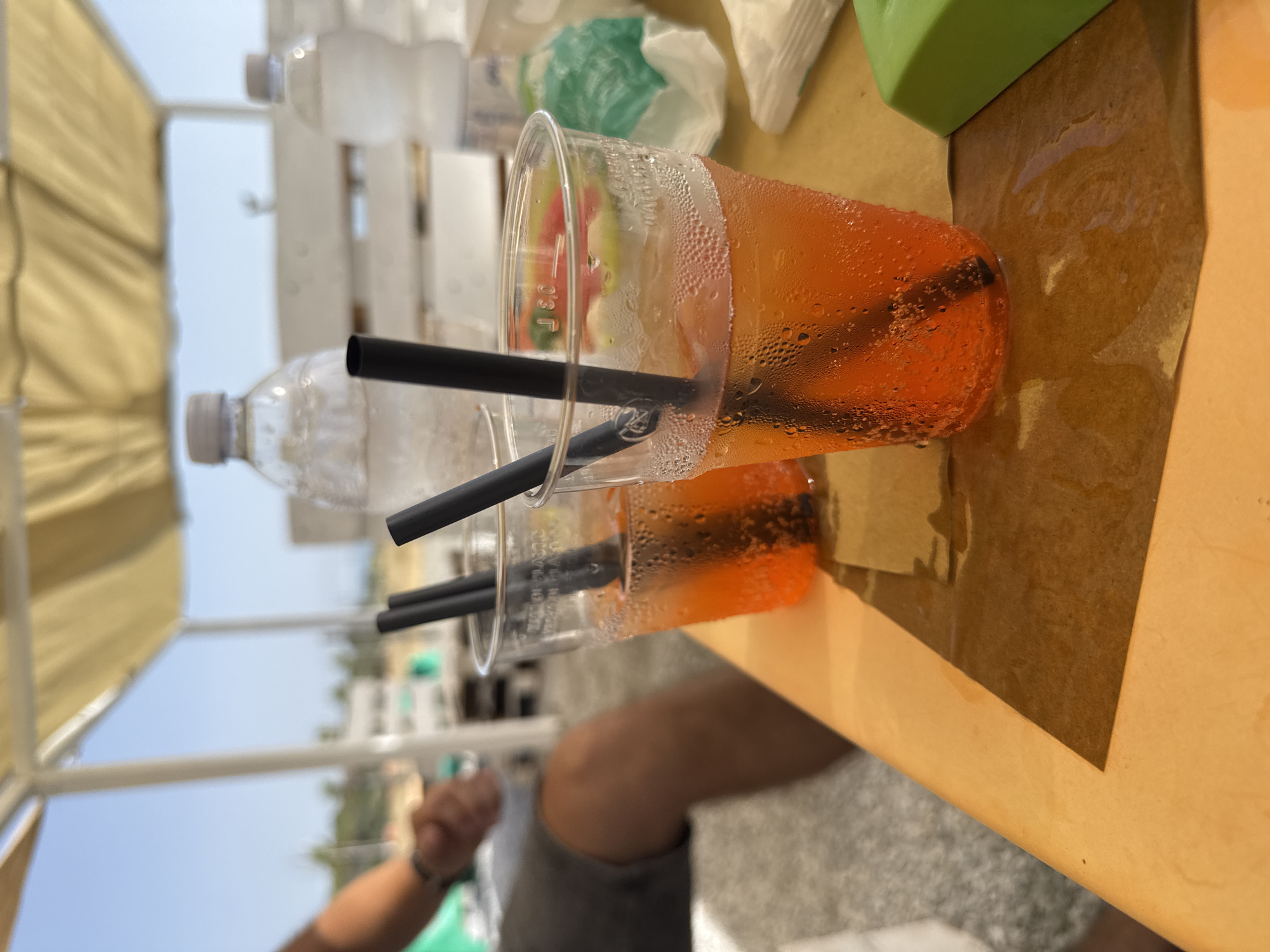 A close-up, eye-level shot captures two clear plastic cups filled with an orange beverage and black straws on an orange table at a beach bar in Lamezia Terme, Italy. The cups are covered in condensation. In the background, a person's legs are visible, clad in grey shorts. Further in the background, several plastic bottles of water and a green object are on display. The scene is illuminated by bright daylight, with a blue sky visible above, suggesting a warm, sunny day at the beach.