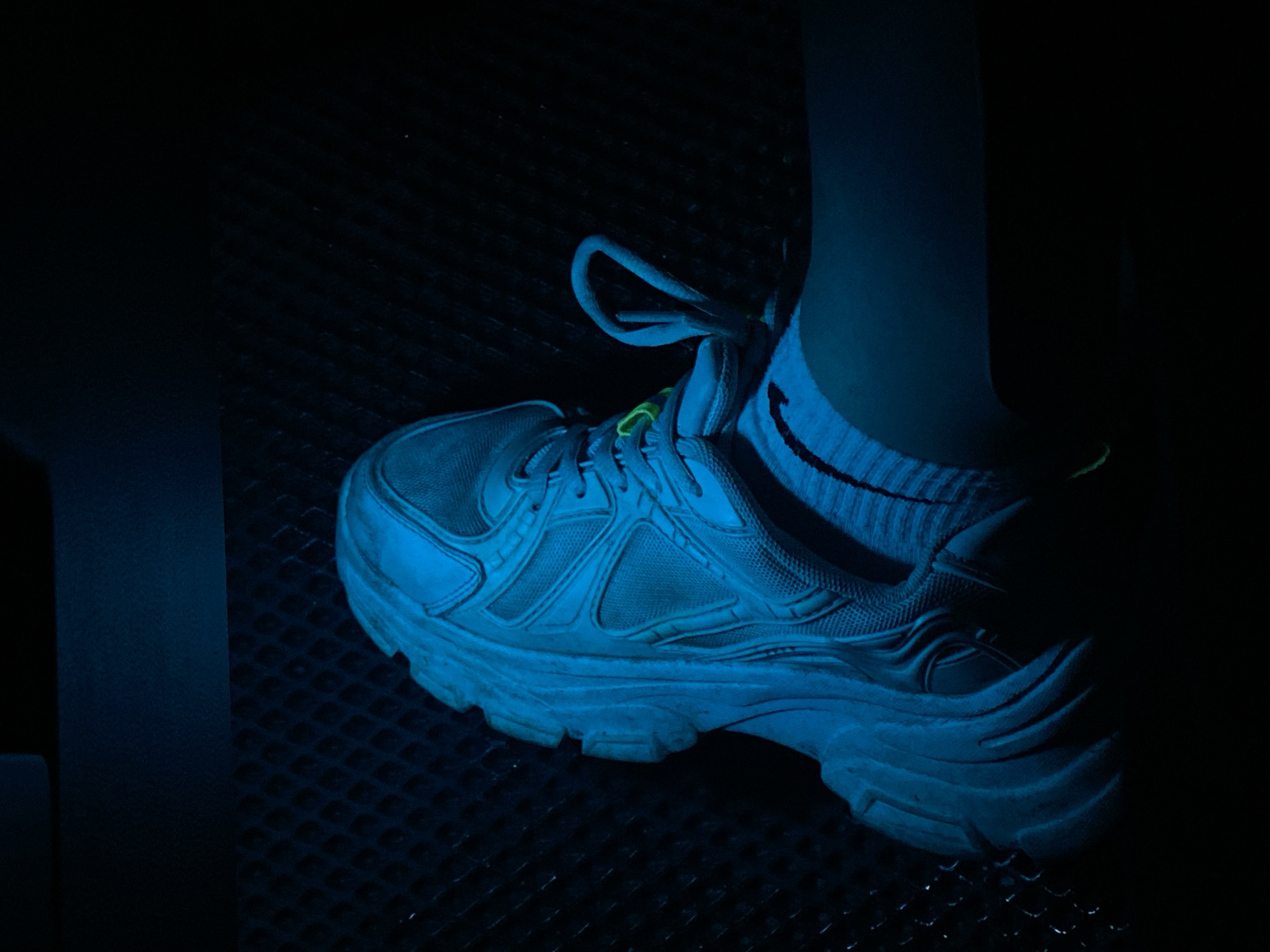 The image shows a close-up view of a person's left foot wearing a white athletic shoe and a striped sock. The shoe has a complex design with mesh panels and a thick sole. A neon green lace loop is visible on the shoe. The foot is resting on a dark, textured surface, possibly a grate or a padded seat. The lighting is predominantly blue, casting a dramatic and somewhat artificial glow on the scene, suggesting it might be under a blacklight or blue stage lighting. The background is very dark, obscuring any details of the surrounding environment. There is no visible indication of the location being in Balashikha, Russia; the image focuses solely on the footwear and a small portion of the immediate surroundings.