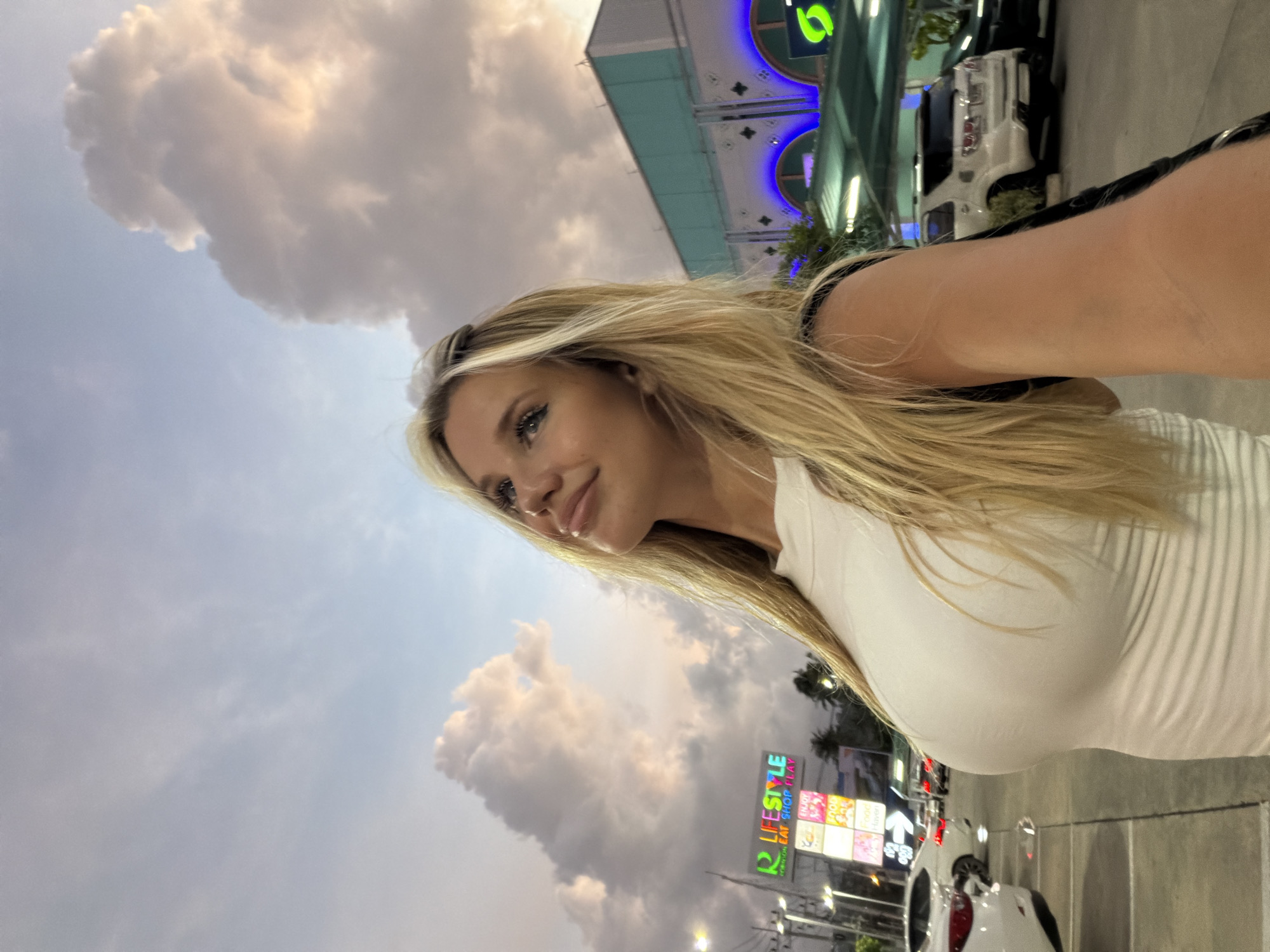 In Si Sunthon, Thailand, a woman with long blonde hair, wearing a white tank top, stands outdoors. She is positioned to the right of the frame, looking upwards and to her left. Her face is illuminated by natural light, and she has makeup on her eyes and lips. In the background, there is a parking lot with several cars, including a white SUV and a white car. A sign that reads "R LIFESTYLE EAT SHOP PLAY" is visible, along with other signs and the facade of a building with blue arched lights. The sky above is filled with dramatic clouds, suggesting it might be evening or during a weather event. The overall scene appears to be a street or plaza setting in a commercial area.