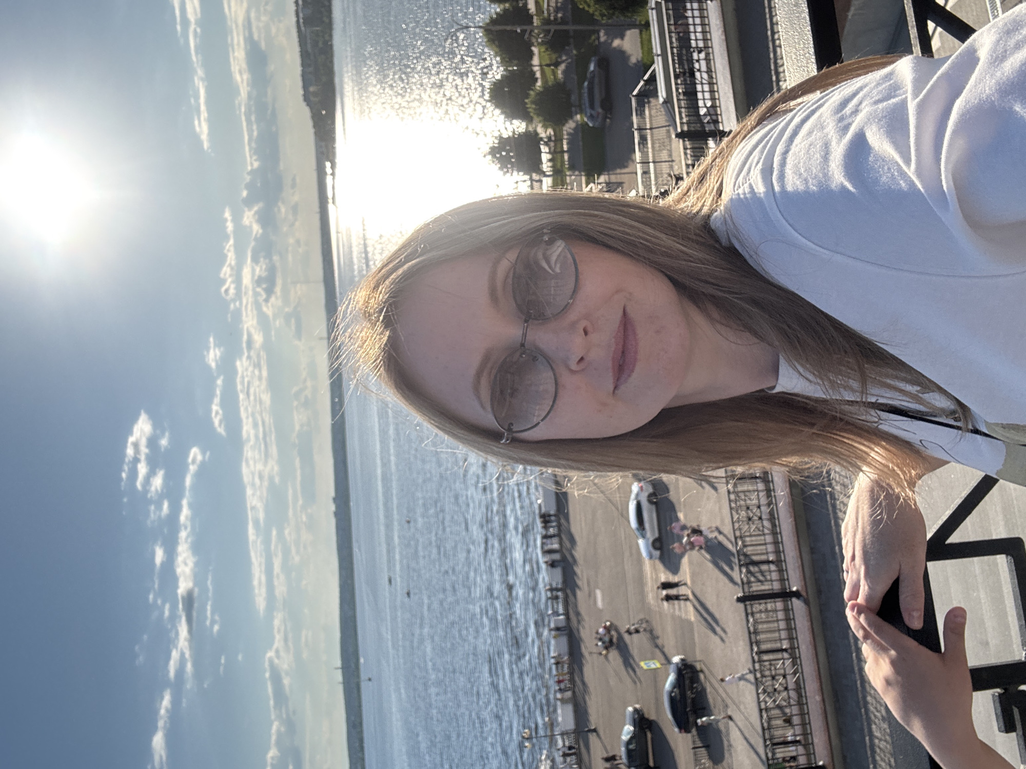 A woman, approximately 30-40 years old, with long light brown hair, is depicted in a sunlit outdoor selfie, wearing dark oval sunglasses and a white long-sleeved top. Her left hand is resting on a black metal railing, with a smaller, juvenile hand also resting on the railing to her left. The scene is set from an elevated viewpoint overlooking the Izhevsk Pond in Izhevsk, Russia, on a bright, sunny day with a clear blue sky and scattered white clouds. The sun, positioned high in the sky, creates intense glare and reflection on the water's surface, particularly on the right side of the image. Several small boats are visible on the water. Below the viewpoint, an urban embankment features a promenade along the water's edge, populated by numerous pedestrians. Further down, a paved road includes various vehicles: a dark pickup truck, a white sedan, and a dark sedan. Additional pedestrians, a cyclist, and two individuals on a recreational tricycle are visible on the road and crosswalks. Black, ornate metal railings separate the different ground levels of the observation point and the waterfront area.