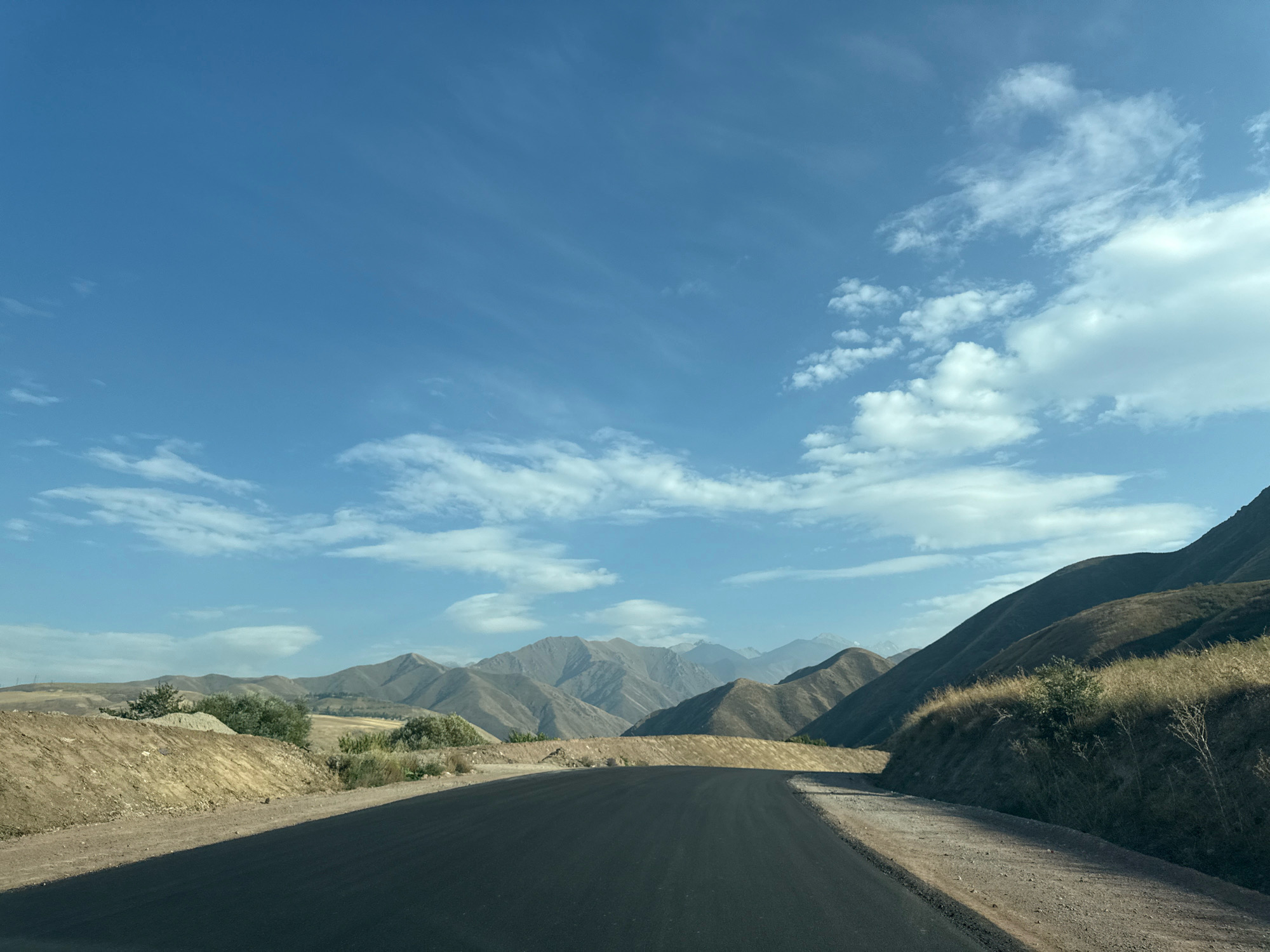 The image shows a curved asphalt road leading through a dry, mountainous landscape. The road is dark gray, with a lighter colored gravel shoulder on the right side. The terrain on either side of the road is arid, with sparse, dry vegetation and brown, rocky soil. In the background, a range of rugged, tan-colored mountains stretches across the horizon, their slopes casting shadows under a bright blue sky dotted with wispy white clouds. The overall impression is one of a remote, undeveloped area, possibly in a mountainous region of Arashan, Kyrgyzstan. There are no people or animals visible in the image.