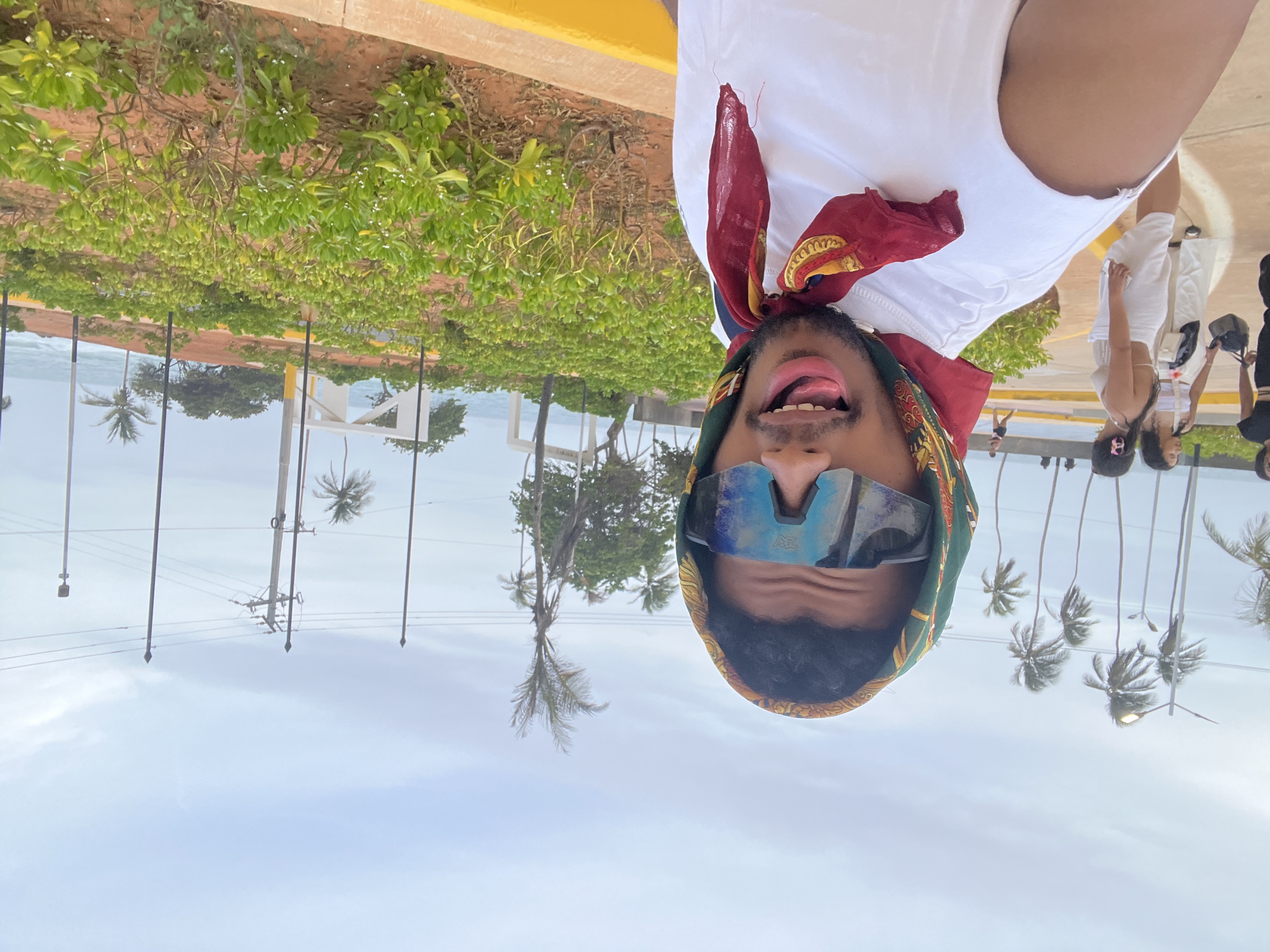 A man with a green and gold headscarf, sunglasses, and a red scarf is the focal point, taking a selfie at Playa El Agua, Venezuela. He is wearing a white shirt and sticking his tongue out. Behind him, there are several people, mostly women, some dressed in white, and palm trees are visible. Overhead power lines and the sky are also present. The environment includes greenery near a sidewalk and what appears to be a beach area.
