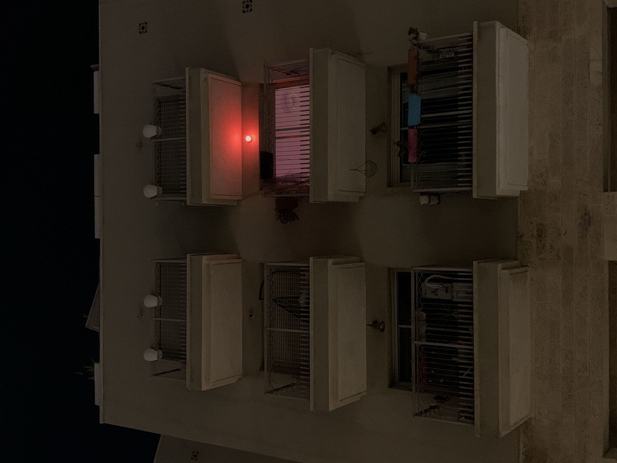 This appears to be an image of the exterior wall of an apartment building in Tel-Aviv, Israel, possibly taken at night. The wall is white or light-colored. The primary features are multiple windows or window openings, most of which have white-painted protective enclosures or shutters. Some of these are open, revealing dark interiors or views through the window. One window has a red light visible inside. Hanging laundry can be seen in the window to the right.