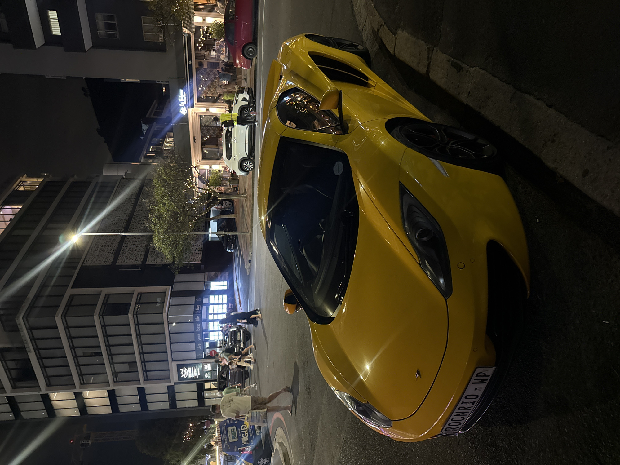 The image depicts a yellow McLaren sports car parked on the side of a street in Cape Town, South Africa, at night. The car's front end is prominently displayed, showcasing its sleek design. The license plate is visible and reads "ROCNR10-WP". In the background, a building with multiple floors and windows can be seen, reflecting the streetlights. A restaurant or cafe with outdoor seating and string lights adorns the street, where several people are walking and socializing. The overall environment suggests a lively urban scene.