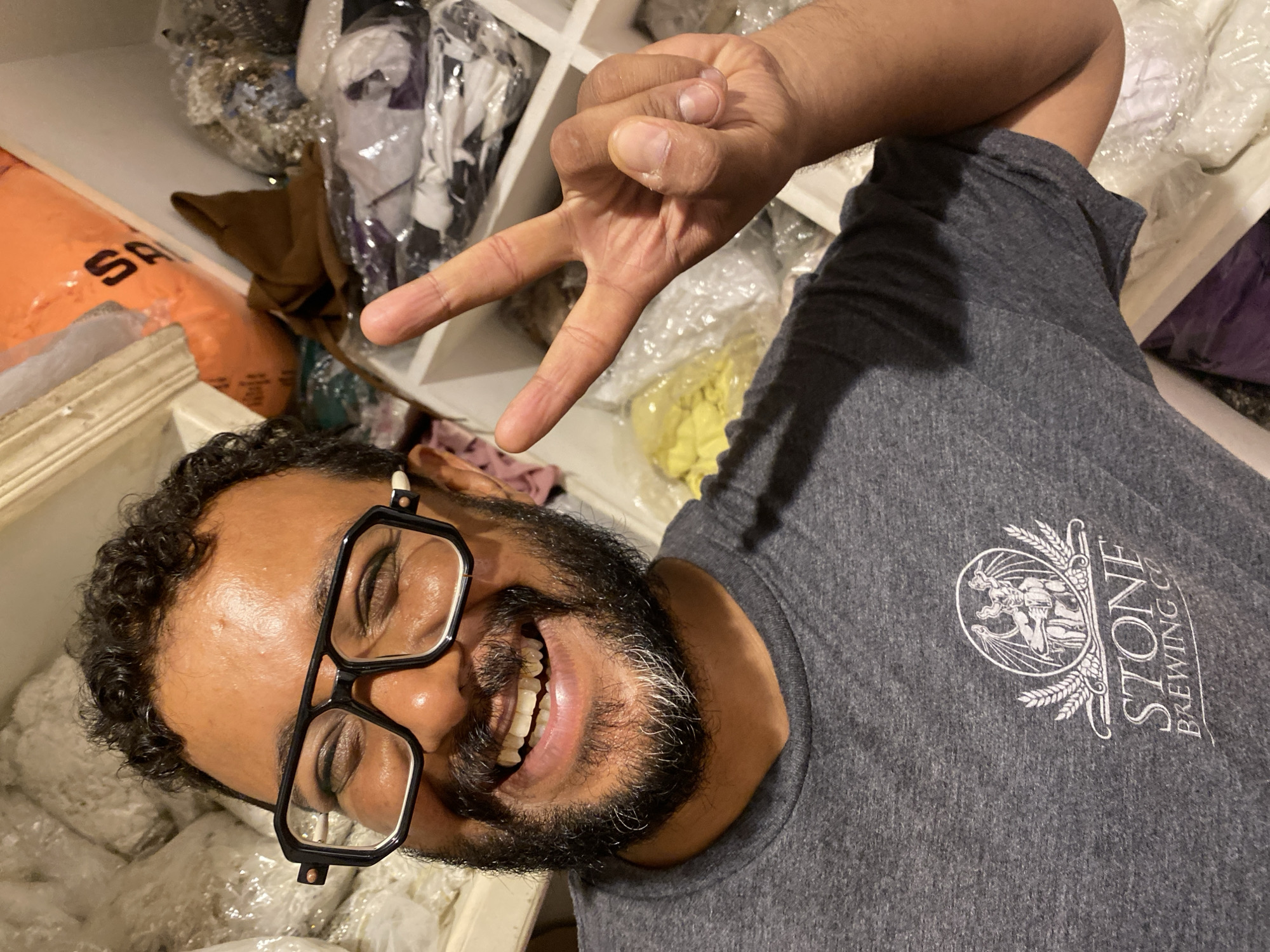 A man with dark curly hair and a beard, wearing black-rimmed glasses and a grey t-shirt with a "Stone Brewing Co." logo, smiles at the camera. He is making a peace sign with his right hand. The man appears to be in a storage area, with shelves filled with various items, including bags of fabric or material, and folded clothing. The background suggests an indoor environment, possibly a warehouse or a large closet, with shelves and possibly a window or light source visible. The man's t-shirt features a circular logo with an illustration of a creature and wheat stalks. The lighting is bright, and the image is a close-up shot focused on the man. No specific location in the city of Legenda, Egypt, can be determined from the visual information provided.