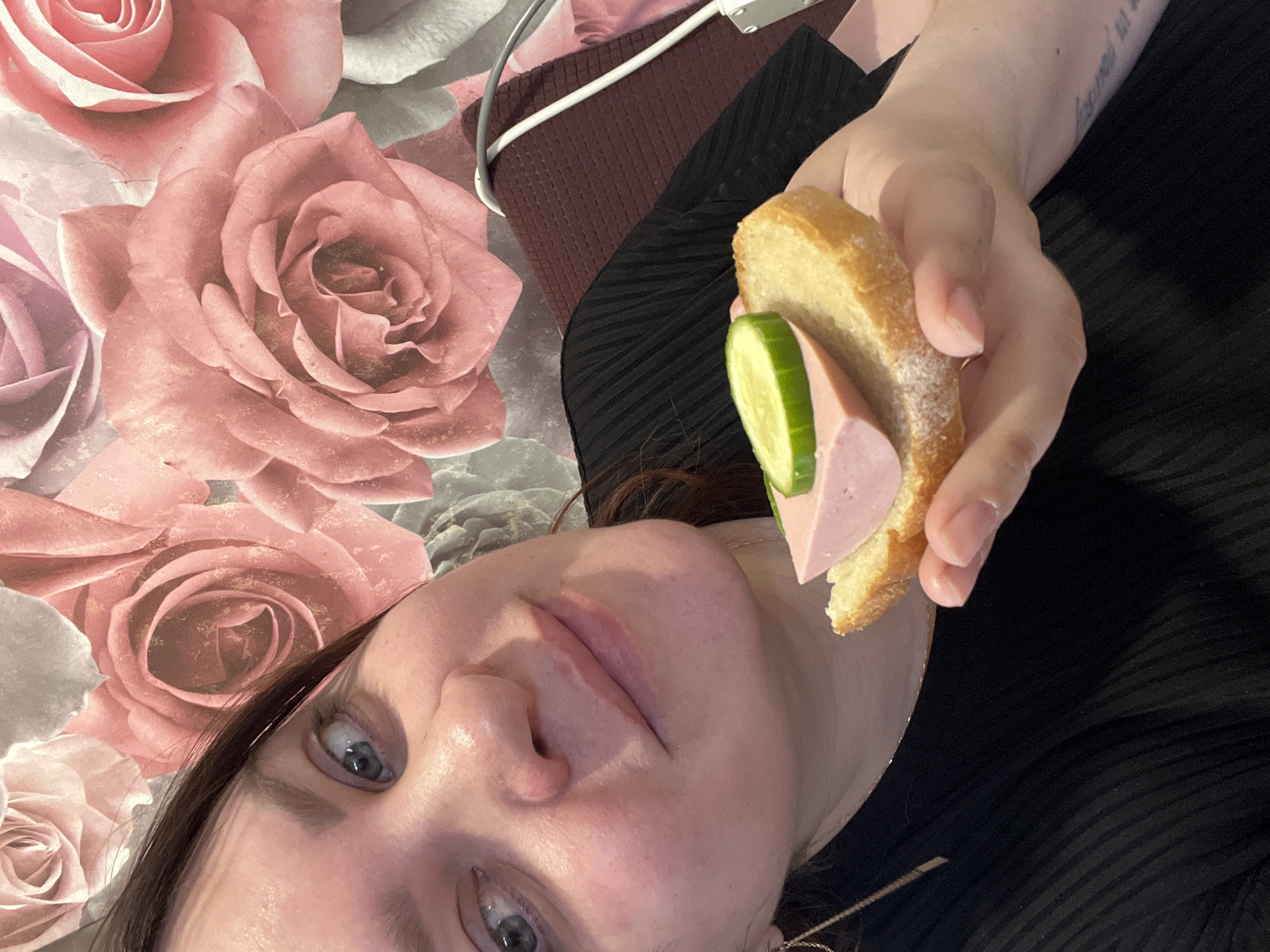 The image depicts a person holding a small sandwich composed of bread, sausage, and cucumber. The background features a floral pattern, and a portion of the person's face is visible. Other items like a cable and a dark-colored garment are also present in the frame. The location is not discernible from the provided image.