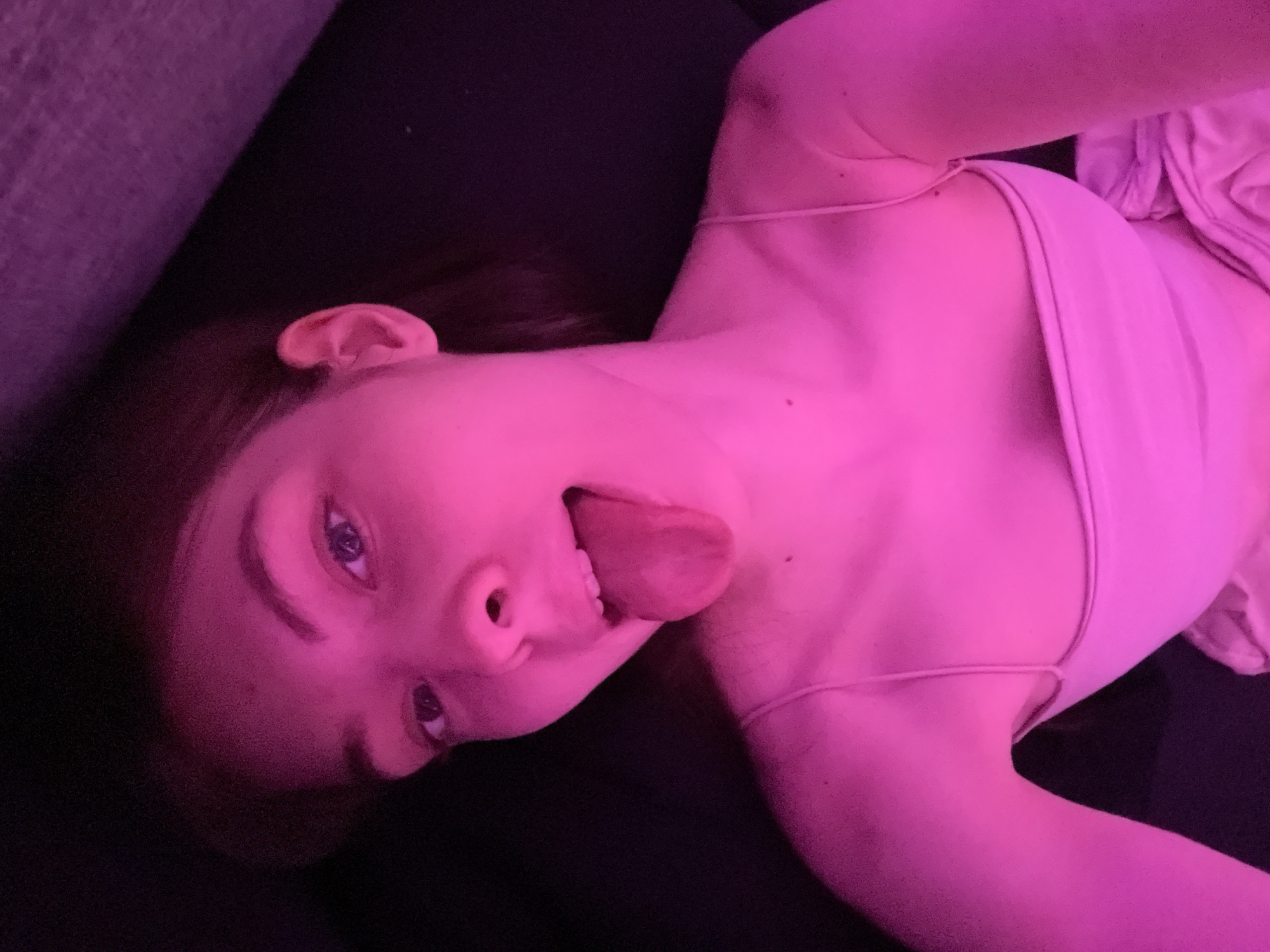 A person lies on a black surface, possibly a couch or bed. The individual has light skin and is sticking their tongue out. They are wearing a light-colored top with thin straps. The scene is lit with a pinkish-purple hue, making it difficult to discern details. The location is not identifiable as Saint Petersburg, Russia, based on the visual information provided.