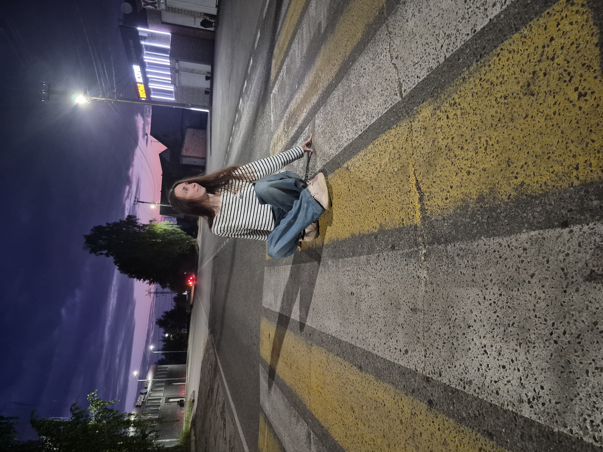 A woman with long brown hair sits cross-legged on a crosswalk during twilight in Chaikovsky, Russia. She is wearing a horizontally striped long-sleeved shirt and loose-fitting jeans. The crosswalk has alternating yellow and white stripes. In the background, buildings with illuminated windows and a street lamp are visible. The sky is a deep purple with hints of pink. A car with its red taillights illuminated is seen driving in the distance. The environment appears to be an urban setting.