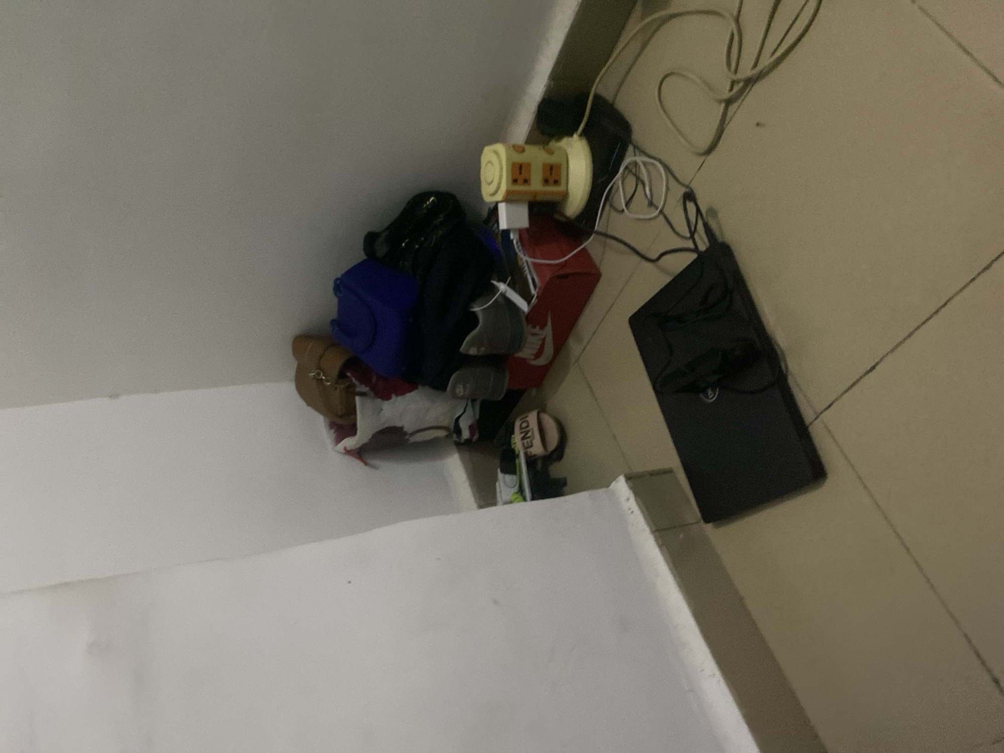 In a corner of a room, a pile of bags and shoes is stacked near a wall. Among the items are a pair of tan sandals with "FENDI" visible on the strap, gray sneakers, and several handbags in blue and brown hues. A red Nike shoebox serves as a base for some of these items. A yellow cylindrical power strip with multiple outlets is plugged in and connected to various cables. To the left, a black laptop is placed on the floor, its lid closed, with a pair of black headphones resting on top. The floor is tiled with a beige color, and the walls are painted white. The scene appears to be in a domestic setting in the city of Howayi, Nigeria.