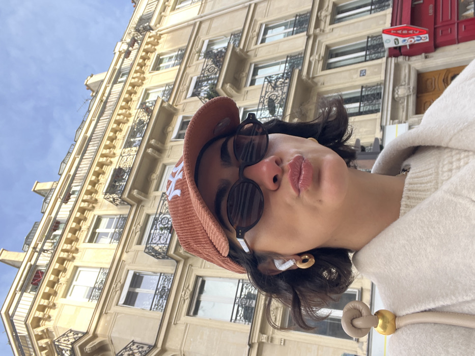 A low-angle, close-up photograph captures a female subject posing on a city street in Paris, France. The subject, positioned centrally, has short dark hair and wears dark oval sunglasses, white wireless earbuds, and gold-colored hoop earrings. She is dressed in a rust-orange corduroy baseball cap featuring a white "NY" logo, a light cream-colored knit sweater, and a matching light-toned overcoat. A beige shoulder bag with a distinct golden knot detail on its strap is visible on her right side. Her lips are pursed forward in a distinct pose.
The background consists of a multi-story, light-colored stone building characteristic of Haussmannian architecture, featuring ornate black wrought-iron balconies on multiple floors. At street level, to the right of the subject, a red storefront with the white word "TABAC" and red neon "OUVERT" (Open) is visible. A building number "24" is affixed to the facade. The sky above is clear blue, indicating daytime.
