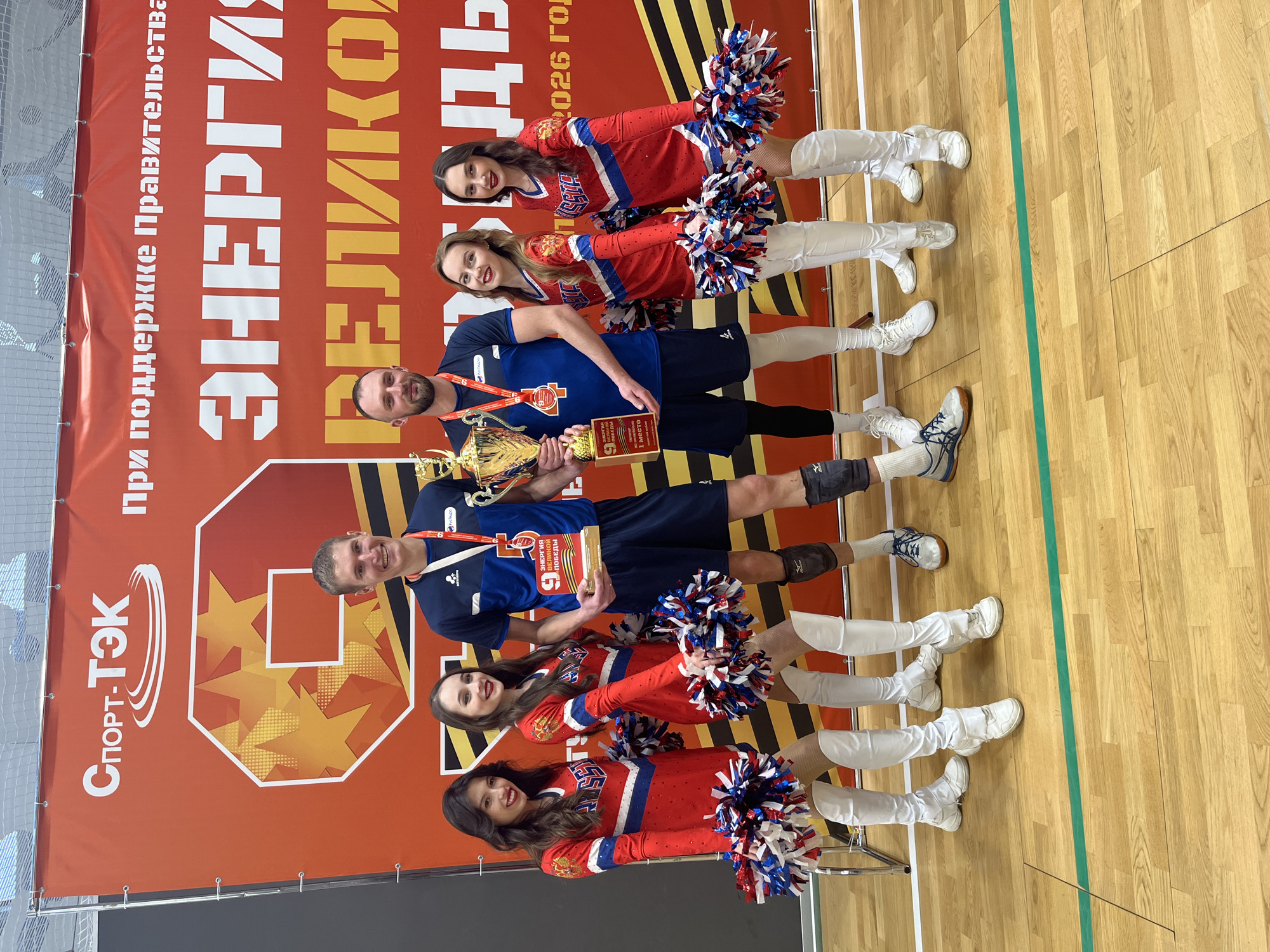 The image depicts a group of people in what appears to be a sports arena or gymnasium. The backdrop is a banner with Russian text including "СПОРТ-ТЭК", "ЭНЕРГИЯ ВЕЛИКОЙ ПОБЕДЫ", and "2026 год". Two men, wearing athletic attire and medals, each hold a trophy and a certificate stating "1 место" (1st place). They are flanked by two pairs of cheerleaders in red and blue outfits with white boots and pom-poms. The floor is wooden with a green line marked on it. The presence of the banner and trophies suggests the scene is a celebration or recognition of a sporting victory, possibly in Moscow, Russia, given the Cyrillic text.