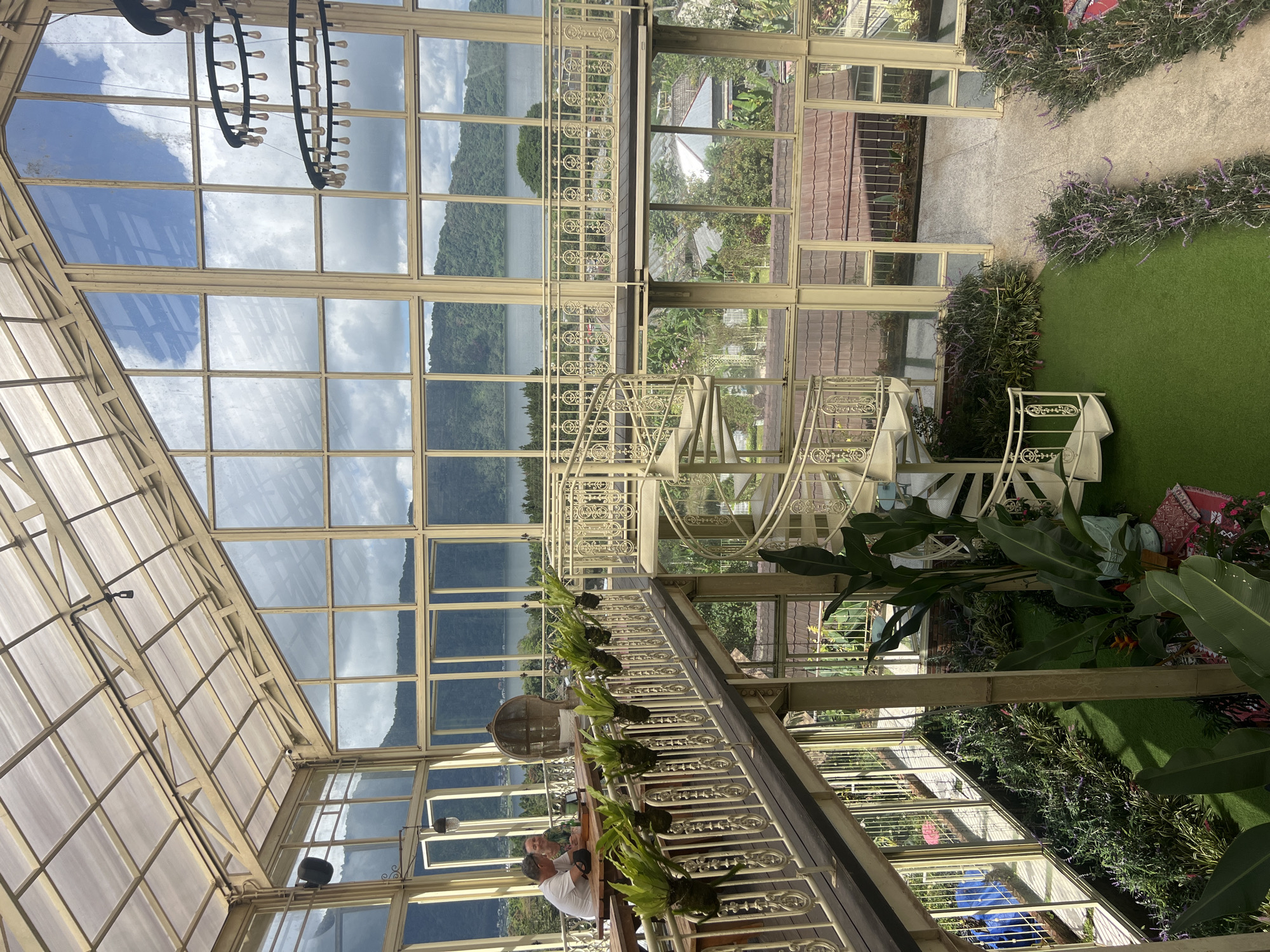 The photograph shows the interior of a greenhouse-like structure, possibly a restaurant or conservatory, in Baturiti, Indonesia. The architecture features a high, latticed glass roof with a chandelier hanging from it. Large glass windows provide panoramic views of a lake and mountains. The interior includes a spiral staircase, ornate railings adorned with plants, and seating arrangements where two men are sitting at a table. The ground level includes greenery and a walkway.