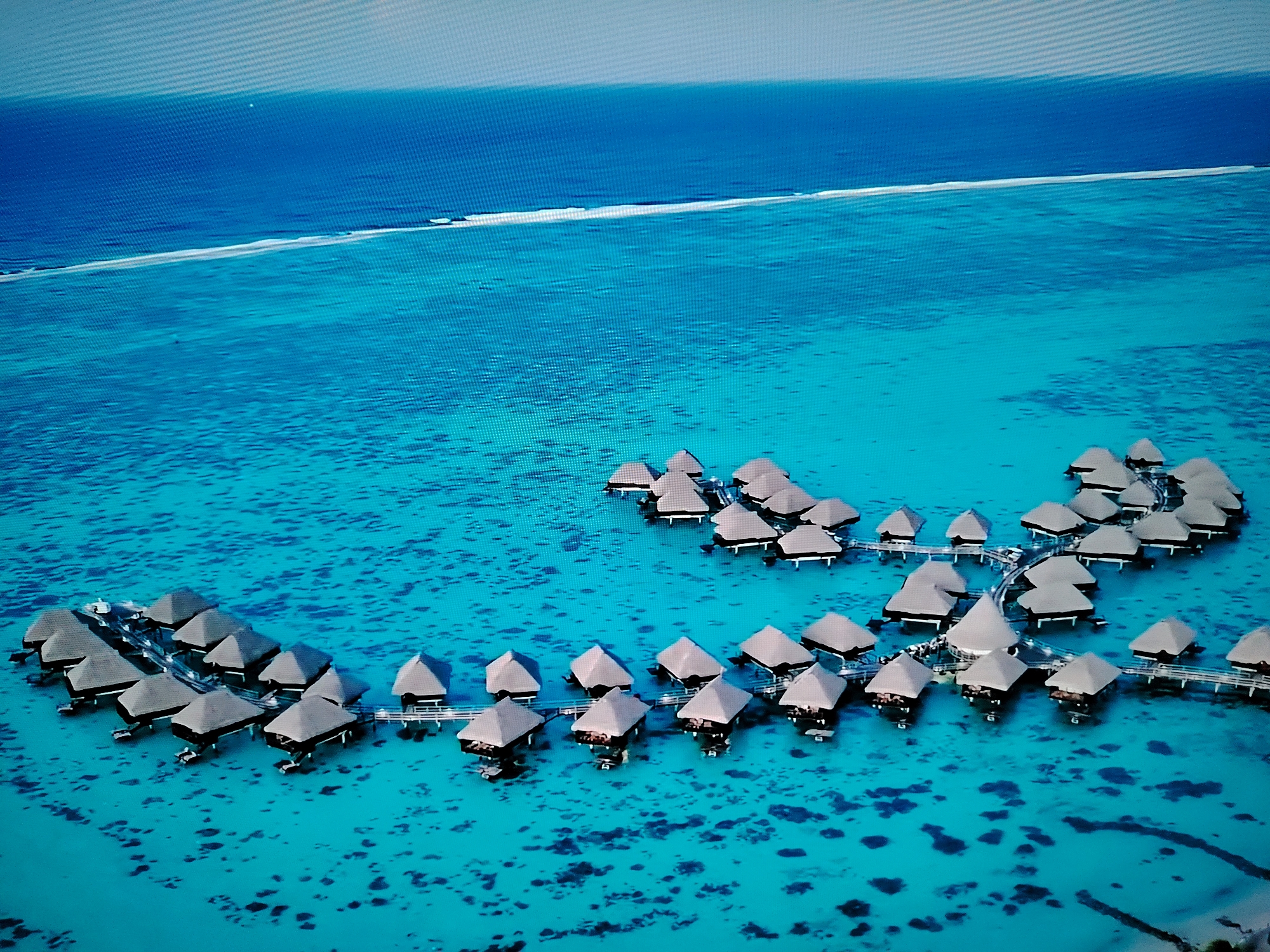 This is an aerial, high-angle, wide shot of an overwater bungalow resort in a tropical, turquoise lagoon. The water is clear, revealing dark coral or seabed formations below. Numerous thatched-roof bungalows are interconnected by wooden walkways, forming clusters. The bungalows are dark gray with lighter-colored roofs and are supported by stilts above the water. In the background, a distinct line of white surf breaks against a sandbar or shallow reef, separating the lagoon from a deep blue ocean. The sky is a light, hazy gray. This scene does not depict Huntingdonshire, United Kingdom, which is a landlocked county.