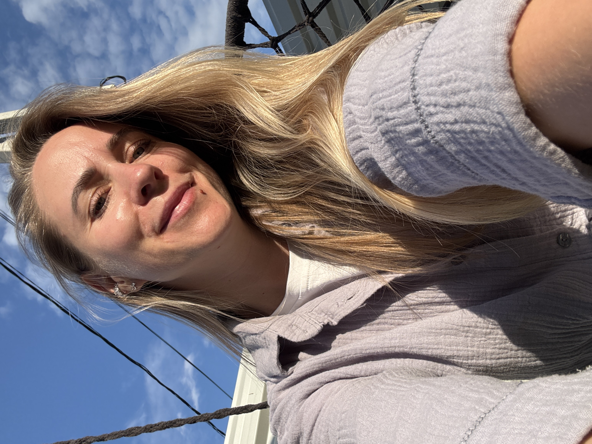 The image is a selfie of a blonde woman outdoors. She is wearing a light purple collared shirt over a white t-shirt and has blonde hair that is partially covering her face. She is smiling and looking at the camera, with sunlight hitting her face. The background consists of a bright blue sky with some scattered clouds, and dark cables or ropes are visible in the foreground and background. The overall lighting suggests it is daytime. The specific location in Chaikovsky, Russia cannot be determined from the image alone.
