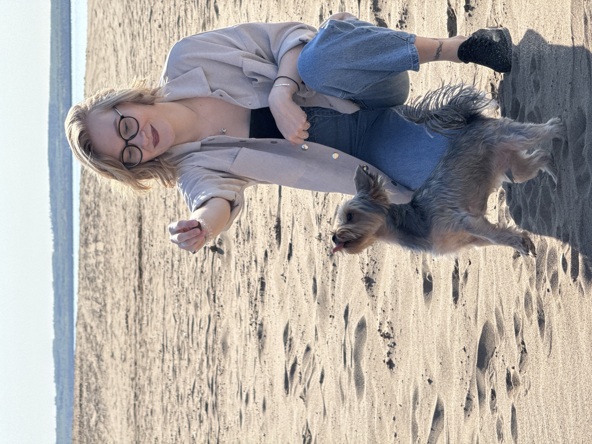 A fair-skinned person wearing glasses, a beige button-down shirt, black top, and blue jeans is lying on the sand in Noviy, Russia. A small, gray-brown Yorkshire Terrier dog is standing next to the person, tongue visible. The person is holding out a hand toward the dog. A sea or lake is visible in the background.