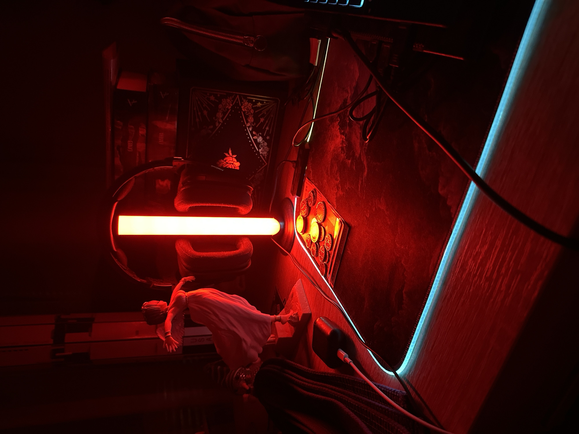 This image depicts an indoor scene, specifically a desk setup in a dimly lit room, located in Unknown, Unknown. The environment is dominated by artificial red lighting from a central, vertical, cylindrical LED lamp positioned on a large, dark mouse pad.
On the left side of the desk, a white classical-style statue of a female figure in a flowing gown, posed with outstretched arms, is illuminated heavily by the red light. To the left of the statue, a dark, folded fabric item is visible, partially obscuring a black rectangular power bank or charging brick with a white USB cable plugged into it.
The central light source, a vertical red LED lamp, illuminates the surrounding area with a strong red hue. In front of this lamp, on the mouse pad, a small, dark tray contains multiple round, metallic objects, possibly coins or tokens, reflecting the red light.
The large mouse pad features an integrated LED strip along its perimeter, emitting a contrasting blue-cyan light. Dark cables are routed across the mouse pad, connecting various devices.
To the right of the central lamp, a dark, square-shaped item with intricate, light-colored patterns, including floral motifs and radiating lines, is present, possibly a decorative box or an item of framed art. Further to the right, a dark bag or backpack with a visible zipper is situated. On the far right, a section of a keyboard with blue backlit keys is visible.
In the background, above the central lamp, a large, dark, curved object, possibly an oversized headset, is present. To the right of this, stacked boxed items, likely retail packaging, are visible, illuminated by the pervasive red light. The overall scene conveys a high-contrast environment due to the dominant red illumination and the contrasting blue-cyan light elements.