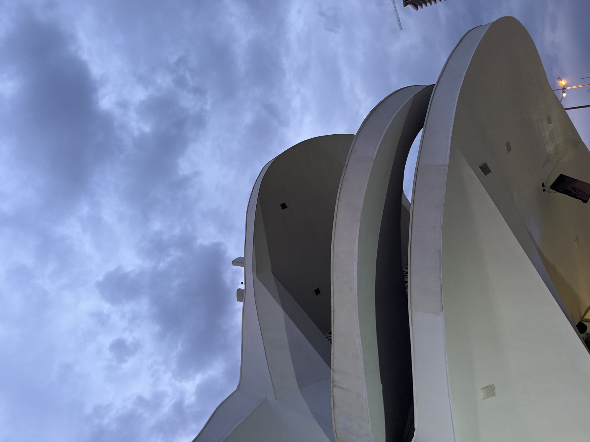 The image captures an upward-angle view of a prominent, modern architectural structure against a cloudy sky. The building is composed of several large, curved panels that are predominantly white or light grey. These panels ascend and appear to overlap or unfold, creating a dynamic, almost wave-like or fin-like silhouette. The design features open, recessed areas between the curves, revealing shadowed interiors.
The sky is densely overcast with dark grey and muted blue clouds, indicating either twilight, dusk, or heavily cloudy weather. The ambient light is low, enhancing the contrast between the illuminated parts of the building and the darker sky. Artificial lights are visible on the structure itself, and a street light or similar fixture with an orange glow is present in the upper right corner, further suggesting a low-light environment. Faint hints of what appear to be construction cranes or tall structures are visible against the sky in the upper left and upper right portions of the frame, hinting at an urban or developing setting.
There are no people visible, and no specific activity or event is taking place, presenting a static architectural study. The focus is entirely on the structure's contemporary design and its interaction with the atmospheric conditions. No visible text is present in the image.