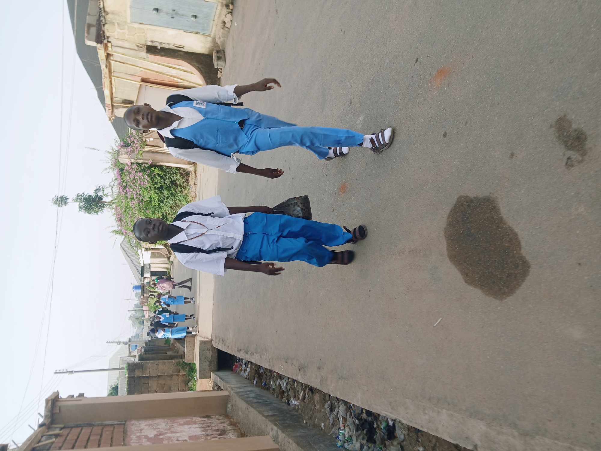 The rotated image depicts a street scene, likely in a developing urban or semi-urban area. The main subjects are two young males, presumably students, walking on a paved road or sidewalk. Both are wearing white long-sleeved shirts and blue trousers. The person further away wears a blue vest over his shirt and white and black sandals. The person closer to the foreground carries a black plastic bag in his left hand and wears brown sandals. Both are looking towards the camera with neutral expressions.
In the background, several other young individuals, also in similar blue and white school uniforms, are visible further down the street, suggesting they are all heading in the same direction or from a school. One person in the distant background appears to be wearing pink.
The setting includes concrete block buildings on both sides of the street, some unpainted or with faded paint, and a few with corrugated metal roofs. A concrete drainage ditch filled with various debris and trash, including plastic bottles and bags, runs alongside the paved surface. Lush green vegetation, including a bush with purple/pink flowers, is visible on the left side. Utility poles and wires are also present in the background.
The ground is a light grey/tan paved surface with some dark stains, possibly from water or oil, and a distinct orange mark. The sky appears bright but hazy or overcast, indicating daytime. There is no visible text in the image. The overall impression is that of students commuting in a community that shows signs of wear and tear, with limited infrastructure and visible litter.