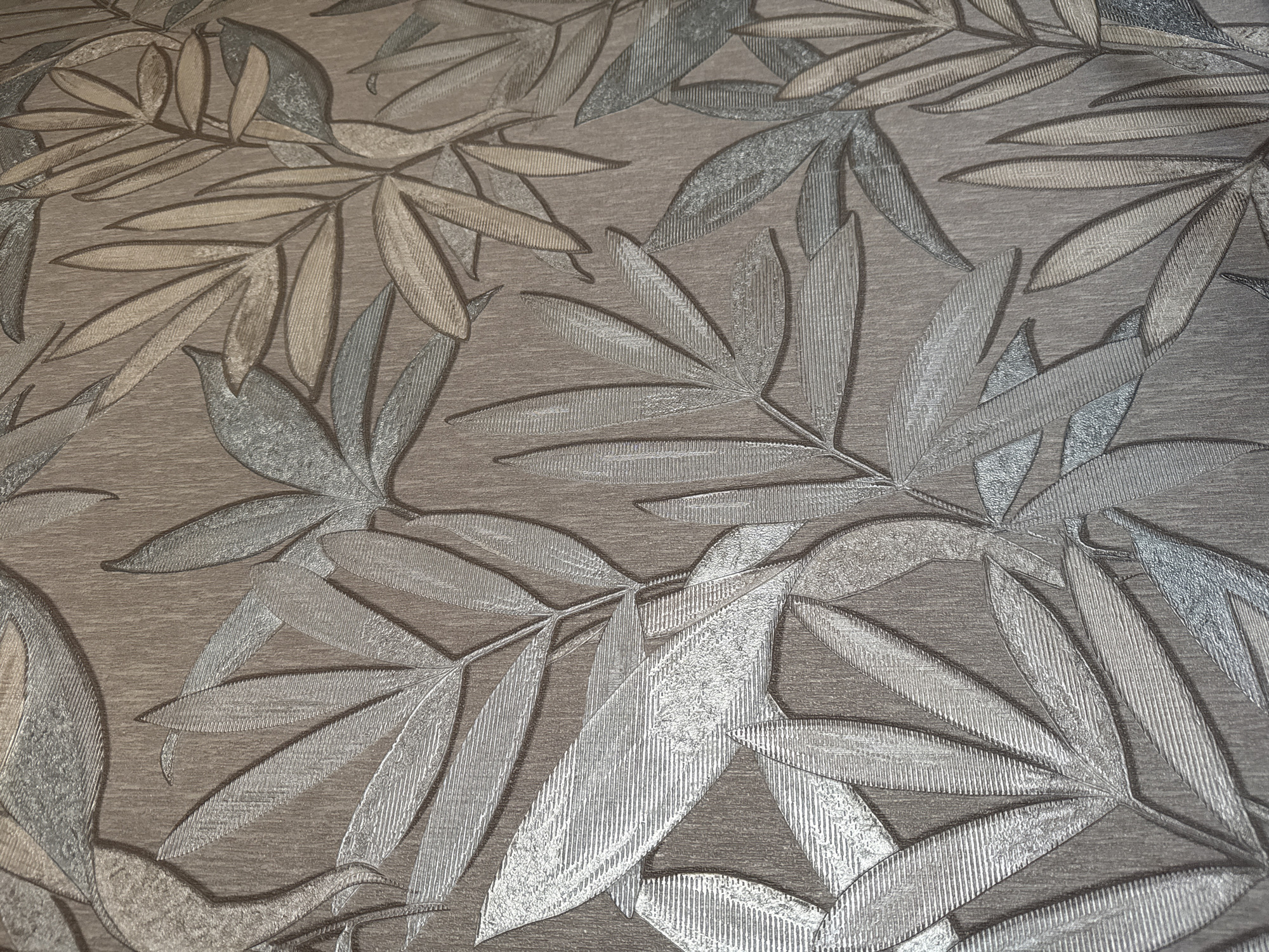 This is a close-up, textured wallpaper pattern featuring stylized leaves and branches. The leaves are elongated and have a ribbed texture, giving them a sense of depth and dimension. The color palette consists of neutral tones, including various shades of brown, beige, silver, and gray, creating a subtle and sophisticated aesthetic. The background also has a textured appearance, resembling wood grain. There are no people, objects, actions, or interactions visible in the image. The scene is a flat surface, likely a wall, adorned with this patterned wallpaper. There is no information about the location in Bishkek City, Kyrgyzstan, provided or discernible from the image itself.