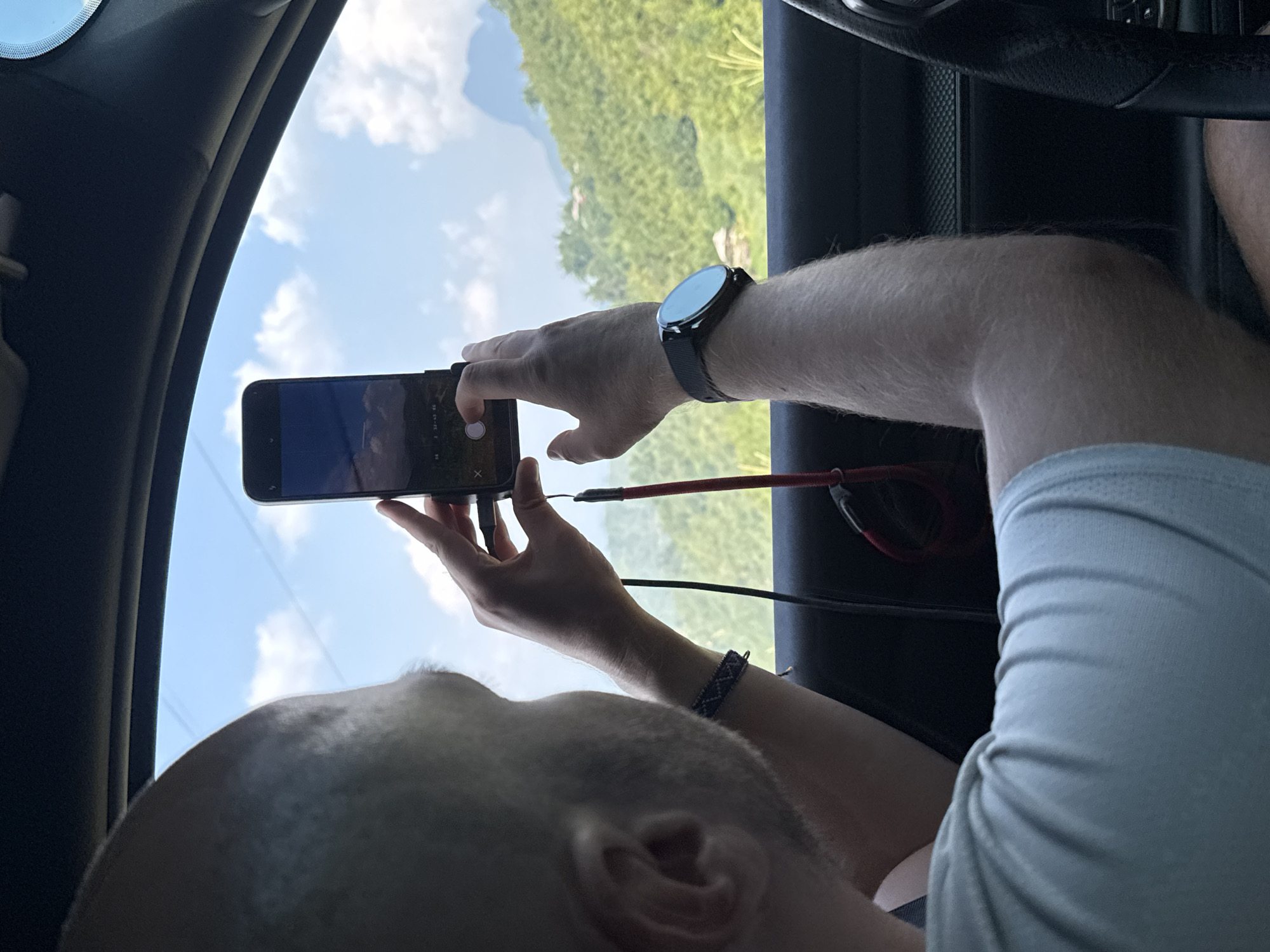 The image shows a person inside a vehicle, possibly a car, taking a photo with their smartphone. The person is male, with a shaved head and fair skin. He is wearing a light-colored shirt and a wristwatch on his left wrist, and a bracelet. The smartphone is black and displays an image. A red and black cable is plugged into the phone. Through the vehicle window, a landscape can be seen, potentially the mountainous terrain found in Mậu Duệ Commune, Vietnam. The presence of a steering wheel indicates the interior is of a car.