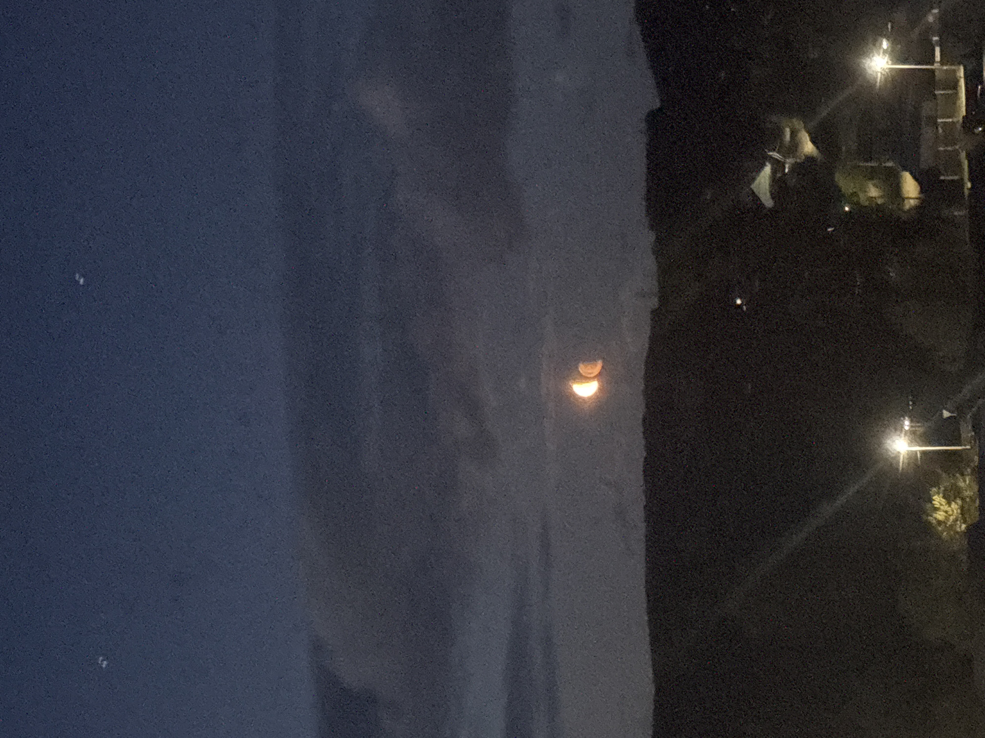 The image depicts a night scene in what appears to be a residential area. The sky is a dark blue, with some clouds. There are two street lights visible on the right, illuminating some trees and buildings. In the center, there is an orange half-moon reflected on the clouds. The image seems to be taken from an elevated position. Given the location (Chaikovsky, Russia) is known, and given that this city is located inland and doesn’t have a coastal area, the reflective image is most likely just clouds, and not water.
