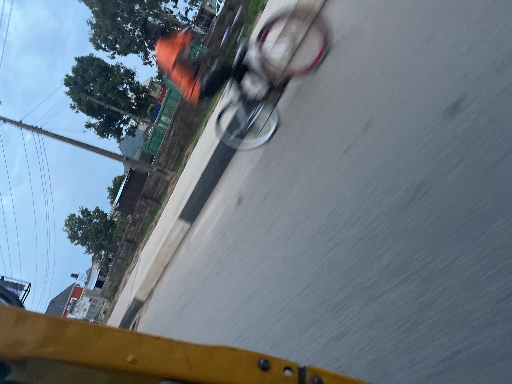 In Bukuru, Nigeria, a person on a bicycle is captured in motion, their form blurred due to the speed of movement. The cyclist is wearing an orange shirt and dark pants. The bicycle itself features both silver and red-rimmed wheels. The scene unfolds on a paved road, bordered by a concrete curb and a grassy embankment. In the background, houses and trees are visible, with power lines stretching across the sky under a cloudy, overcast atmosphere. The foreground is dominated by the yellow panel of what appears to be a three-wheeled vehicle, suggesting the photograph was taken from within or beside it.