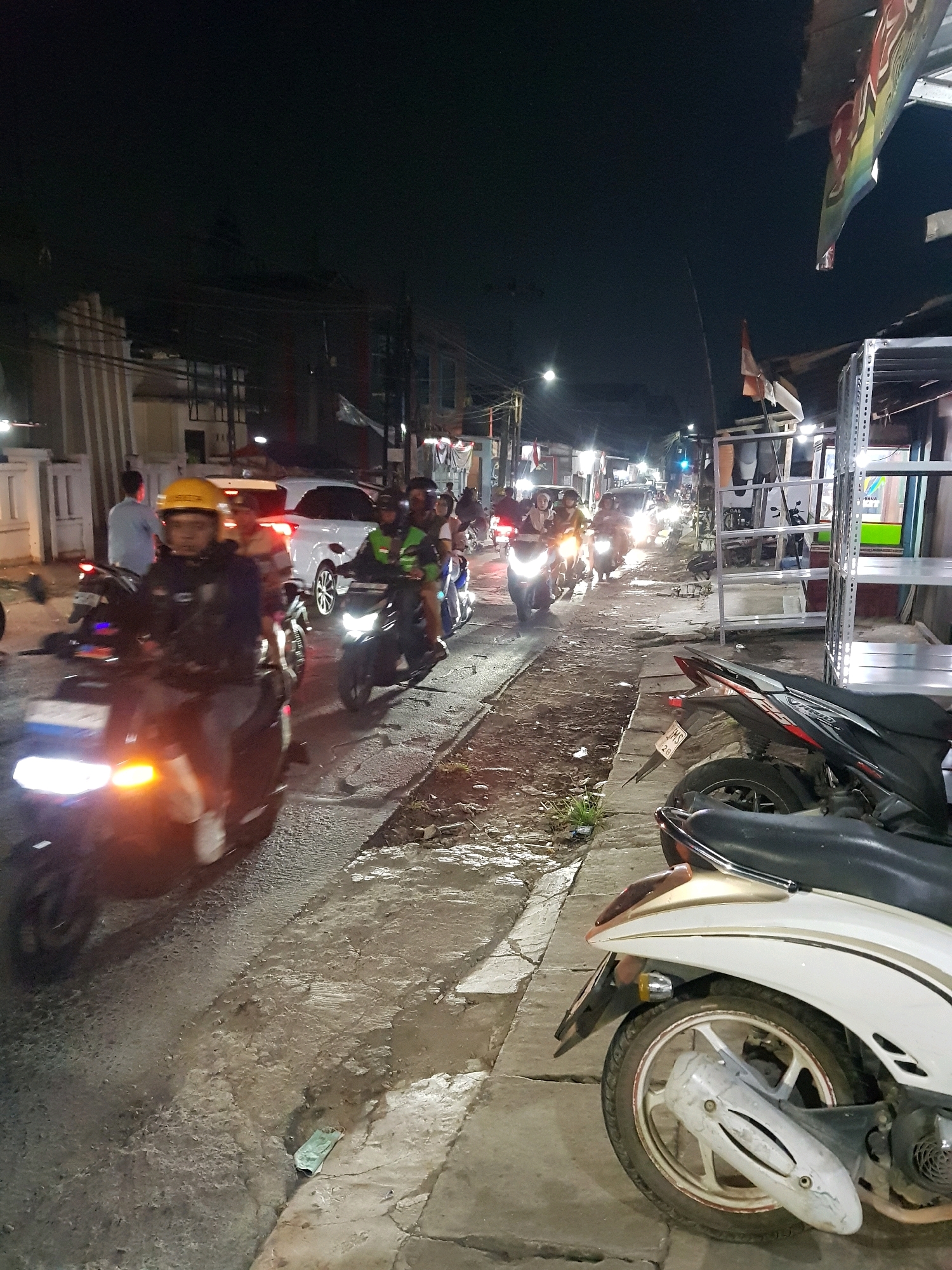 The image captures a nighttime scene on a street in Setiamekar, Indonesia, characterized by moderate traffic and pedestrian activity. Several motorcycles are in motion, with their headlights illuminated, indicating movement along the paved road. One motorcycle in the foreground is ridden by an individual wearing a yellow helmet and a dark jacket. Another motorcycle behind it has a rider with a green vest. In the background, more motorcycles and a white car are visible. The street is lined with buildings and structures, including what appears to be a multi-tiered shelving unit on the right side of the frame, suggestive of a commercial area or market. The ground surface is uneven, with patches of dirt, gravel, and concrete. Streetlights and the lights from vehicles and buildings provide illumination against the dark night sky. The overall atmosphere is one of a busy urban street at night.