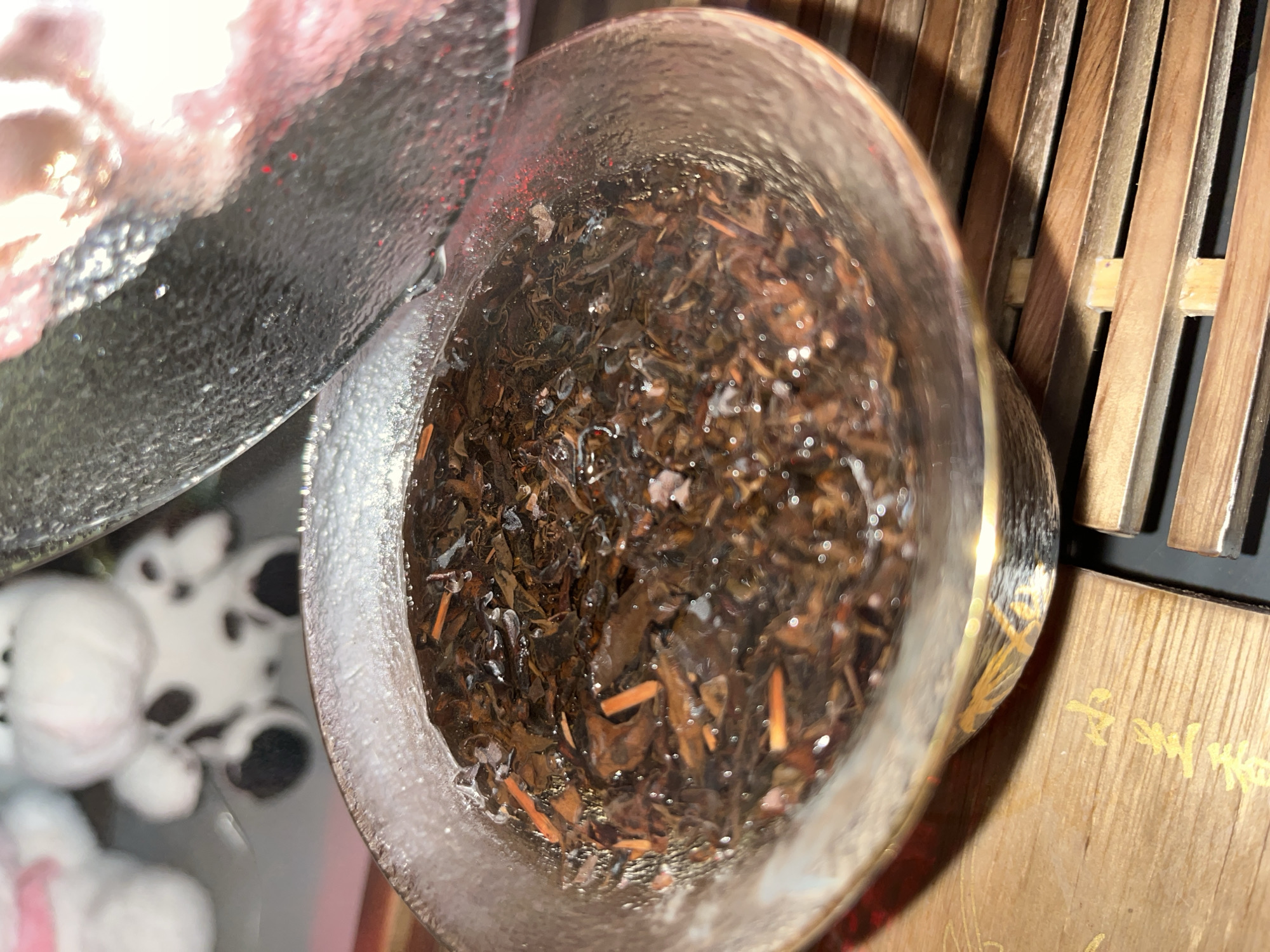 A close-up shot shows a clear glass bowl filled with steeped tea leaves and liquid. The bowl has a gilded rim and sits on a wooden grate. The background is blurred, but a portion of a shiny, reflective lid is visible above the bowl. In the far background, out of focus, are two plush toy animals, one white with black spots and the other predominantly white with a pink ribbon. The scene appears to be indoors, possibly a setting for a tea ceremony, with soft lighting.